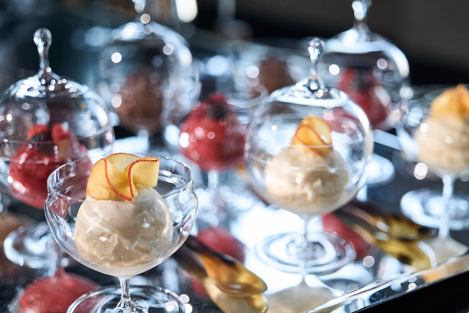 Scoops of ice cream in glass coupes garnished with apple slices, alongside berry sorbets on a mirrored tray