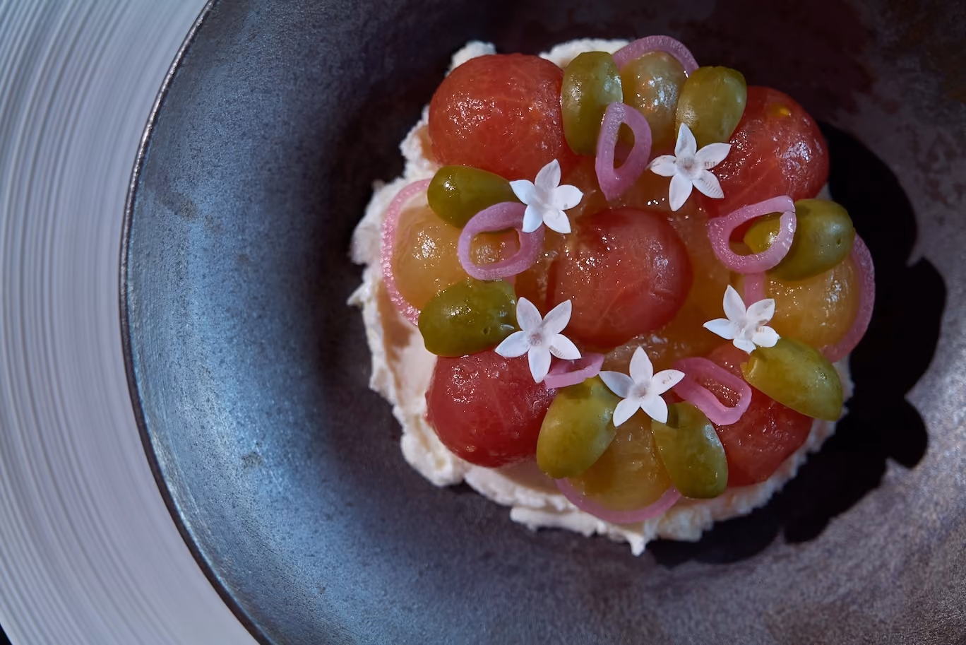 Overhead view of whipped cheese topped with heirloom tomatoes, olives, pickled shallot rings, and white flowers