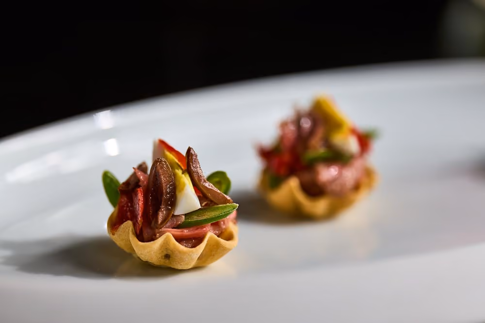 Two tartlet shells topped with cured meat, egg, and olives