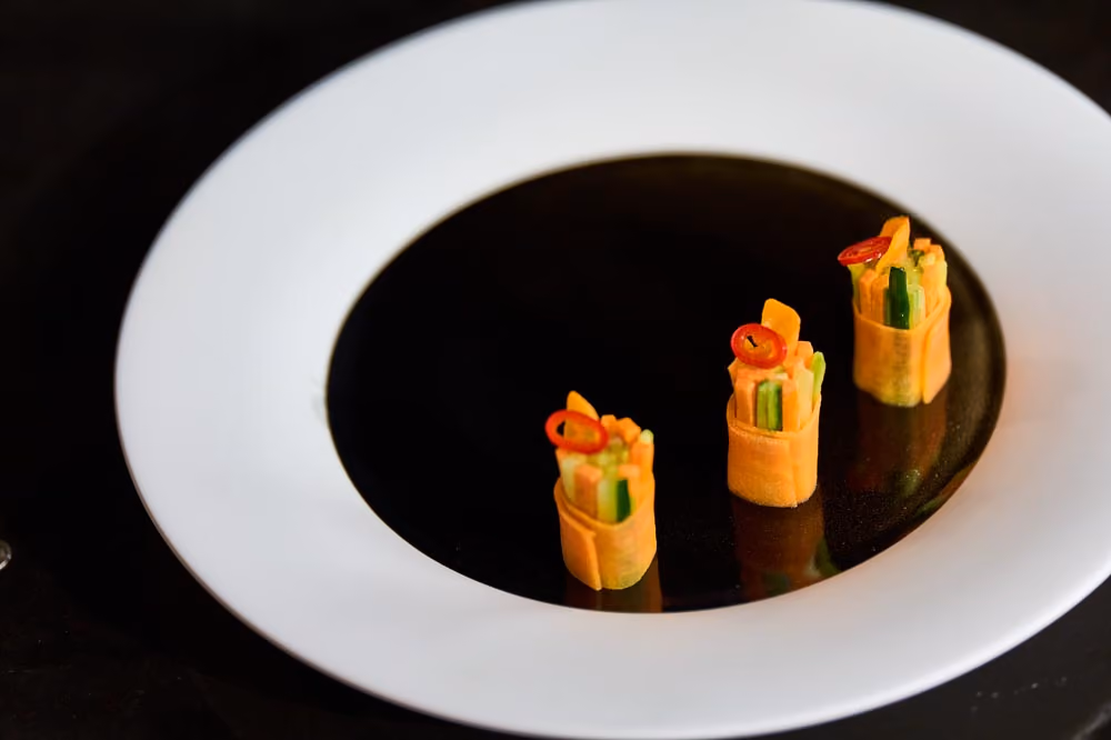 Three vegetable roll canapés wrapped in carrot ribbon and topped with red chili