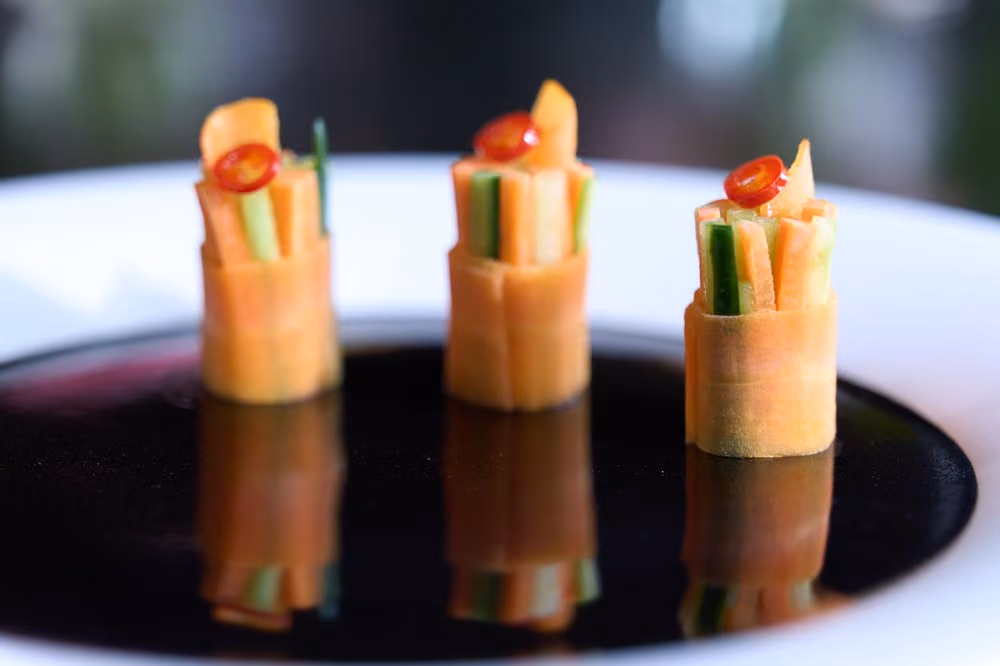 Three vegetable roll canapés wrapped in carrot ribbon and topped with red chili