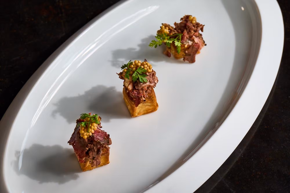 Three canapés topped with sliced beef, mustard seeds, and herbs on a white platter
