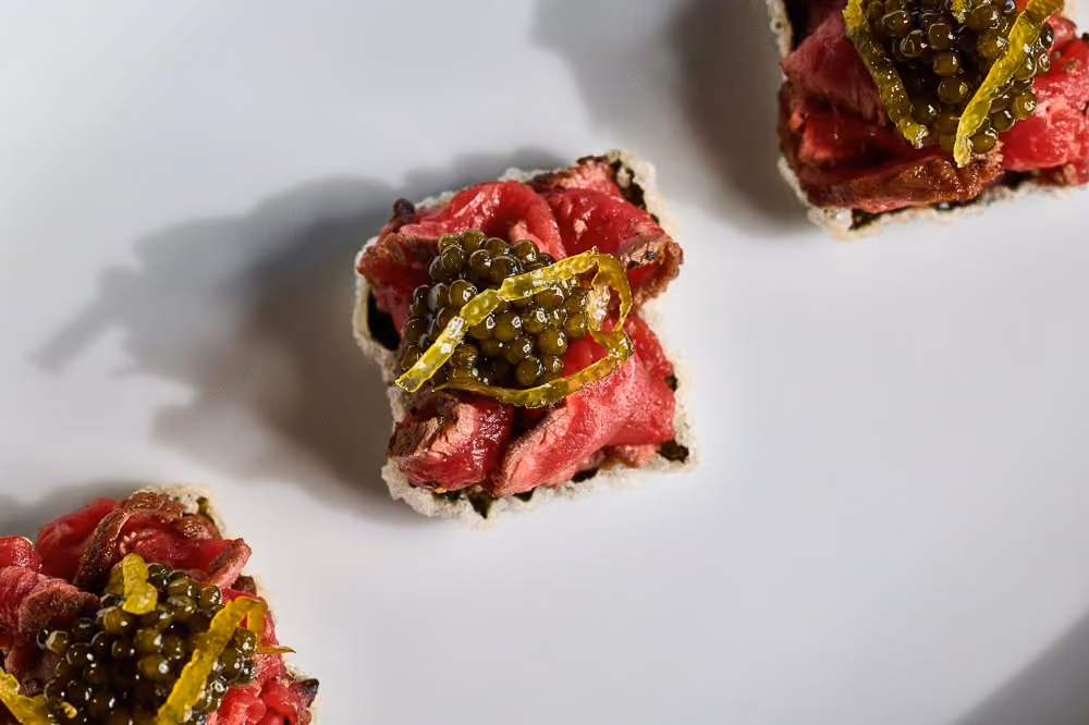 Overhead row of beef canapés topped with caviar and citrus zest on crisped rice cakes