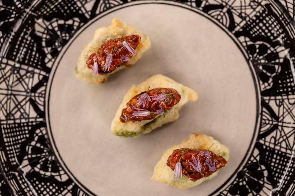 Three crispy canapés topped with tomato compote and purple chive flowers on a patterned plate