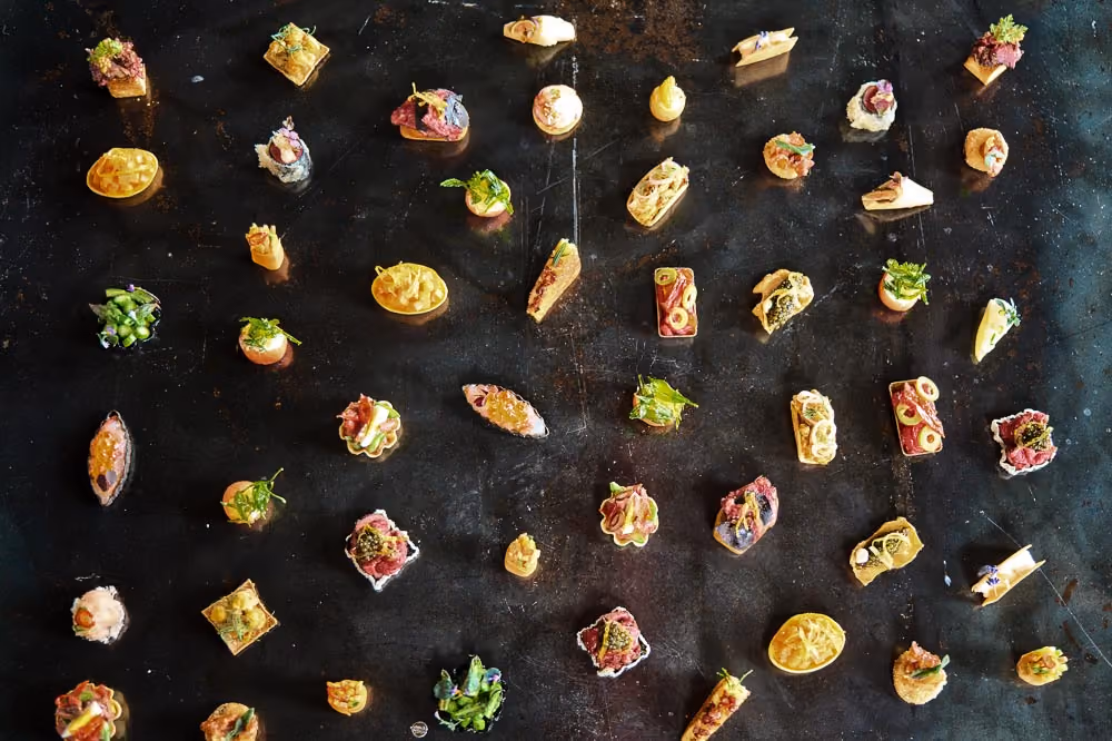 Overhead grid of assorted bite-sized canapés arranged on a dark tray