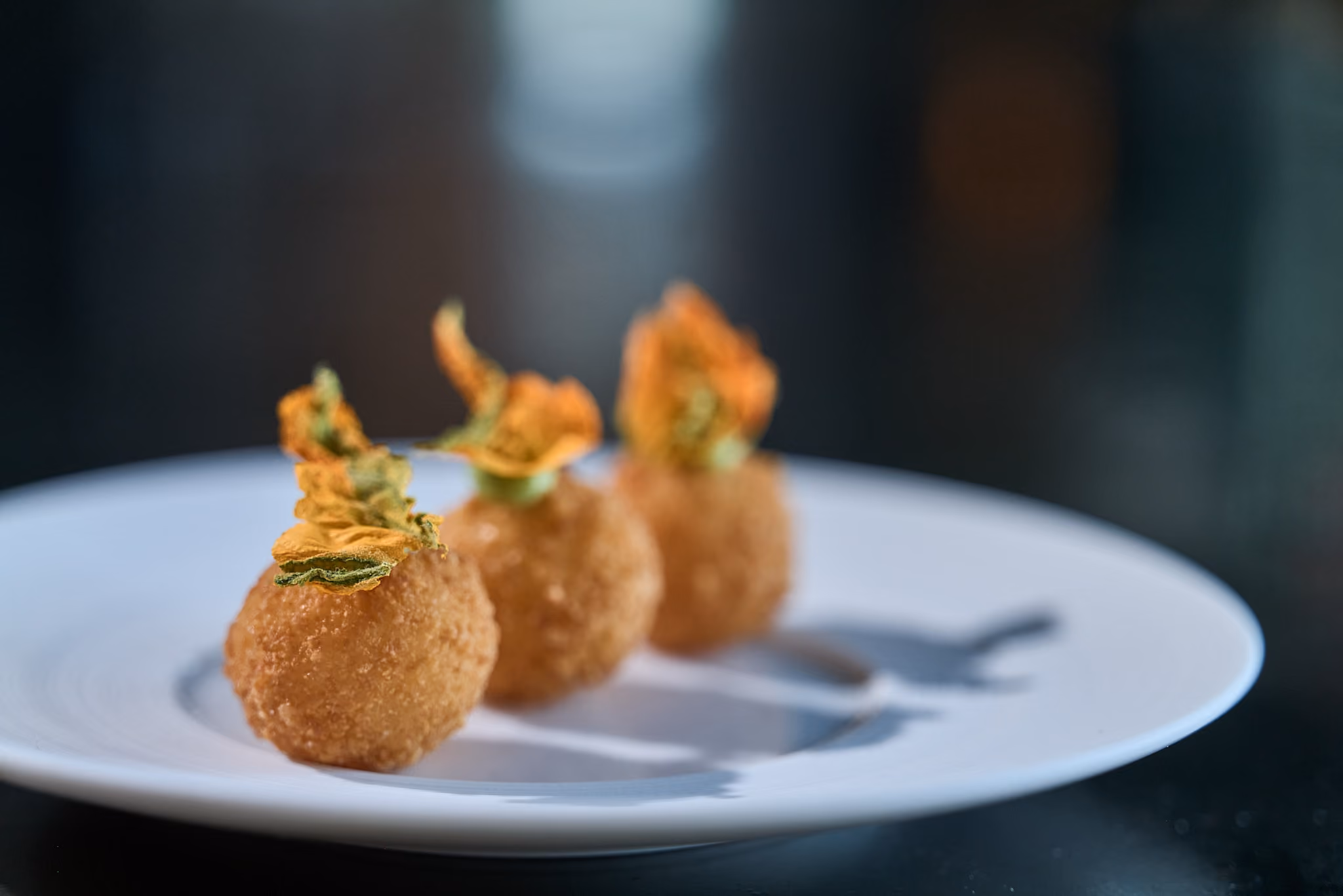 Three fried croquette bites topped with crispy squash blossom on a dark plate