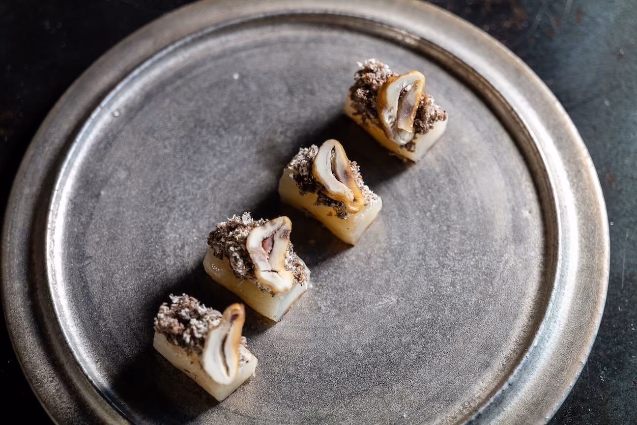Row of rice cake canapés topped with abalone and a crispy garnish on a metal plate
