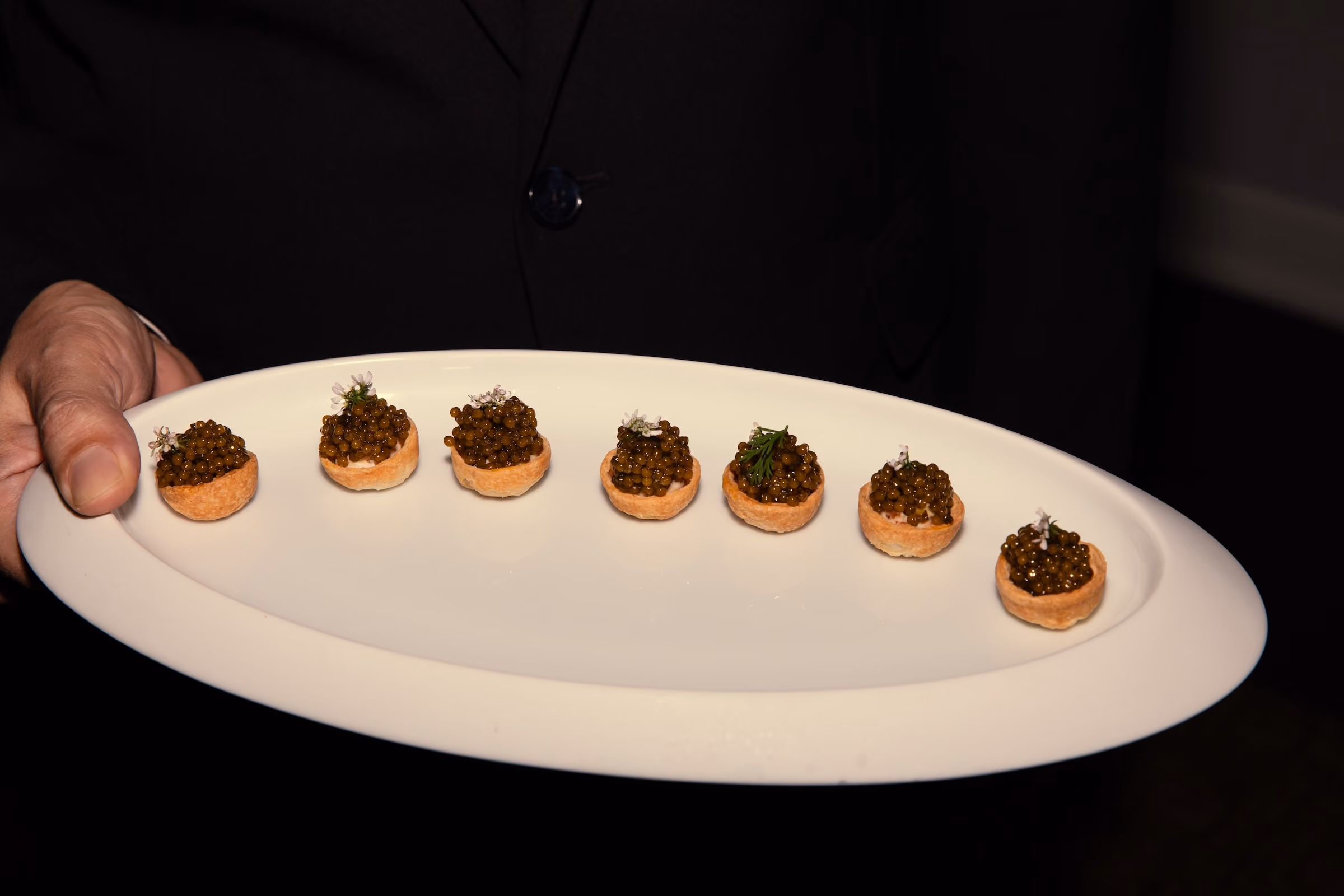 Row of tartlet canapés topped with caviar and flowers on a plate held by a server