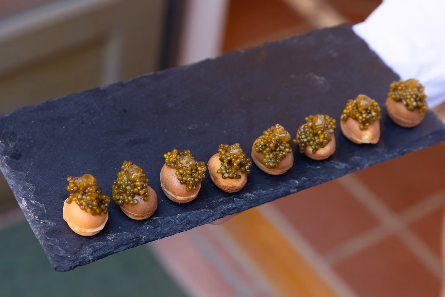 Row of choux pastry canapés topped with golden caviar on a slate board held by a server