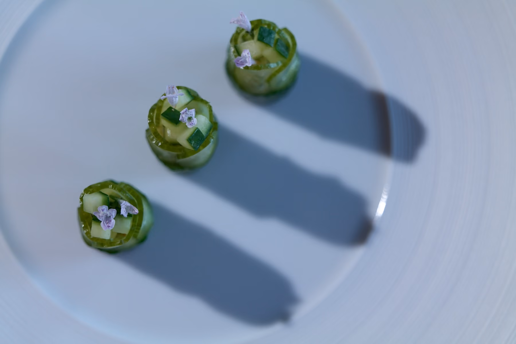 Overhead trio of cucumber roll canapés topped with diced vegetables and purple flowers