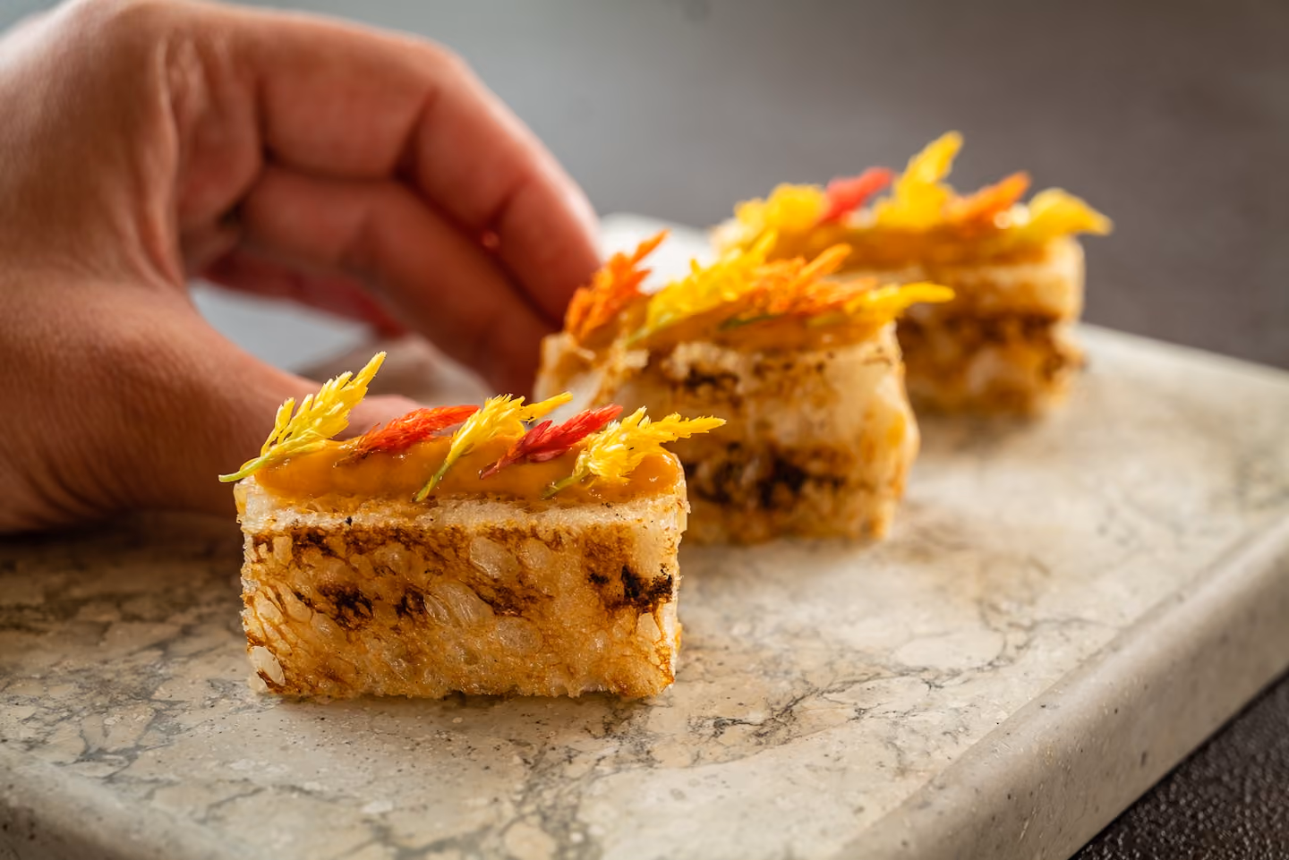 Toasted bread canapés topped with sauce and colorful edible flowers as a hand reaches for one