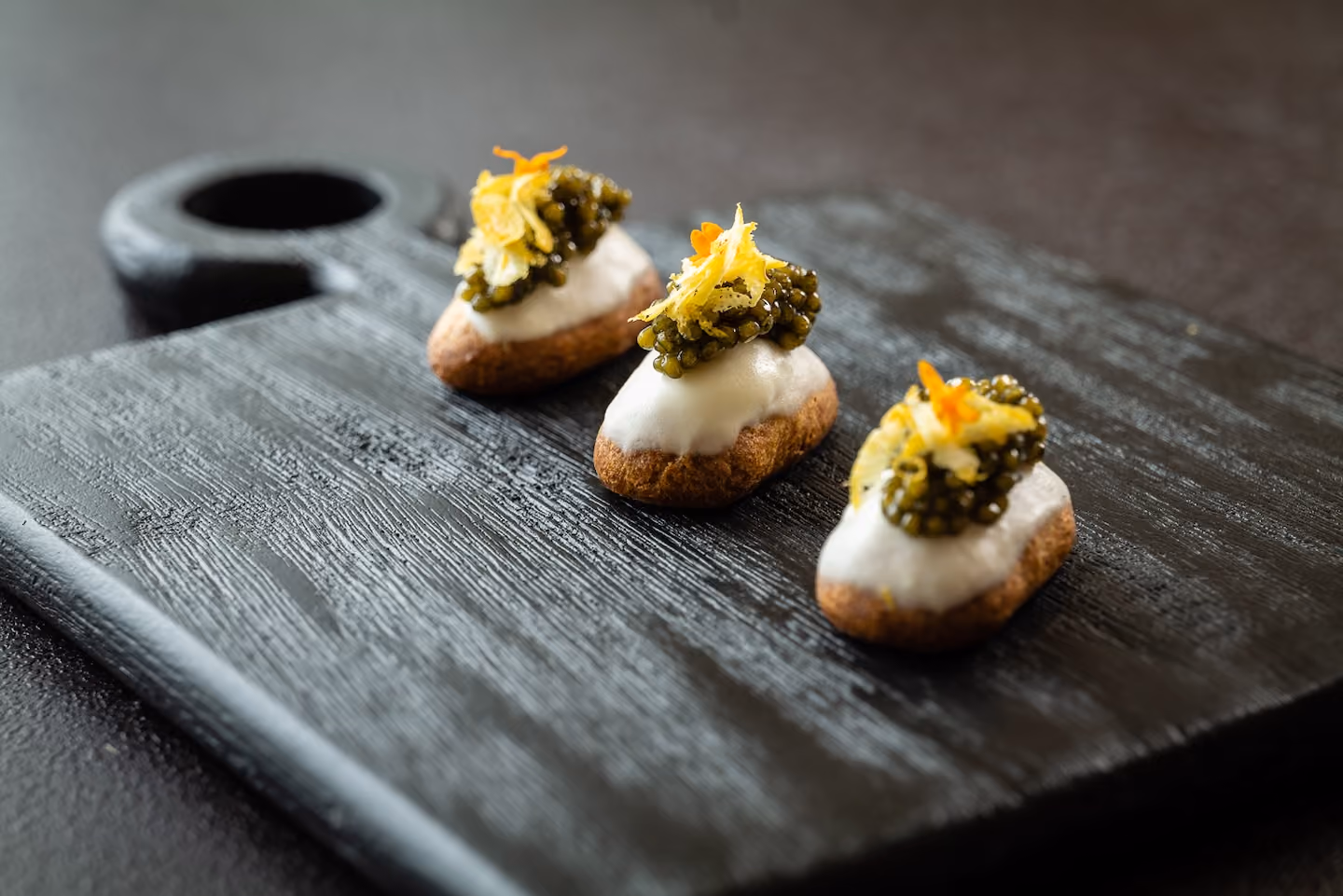 Eclair-shaped canapés topped with caviar and candied citrus zest on a dark board
