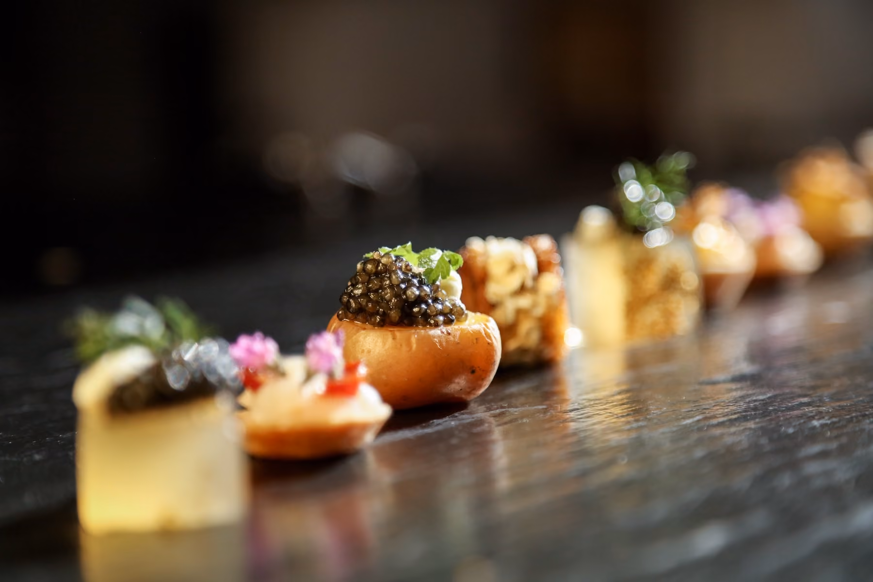 Row of assorted bite-sized canapés with caviar and gold leaf on a dark table