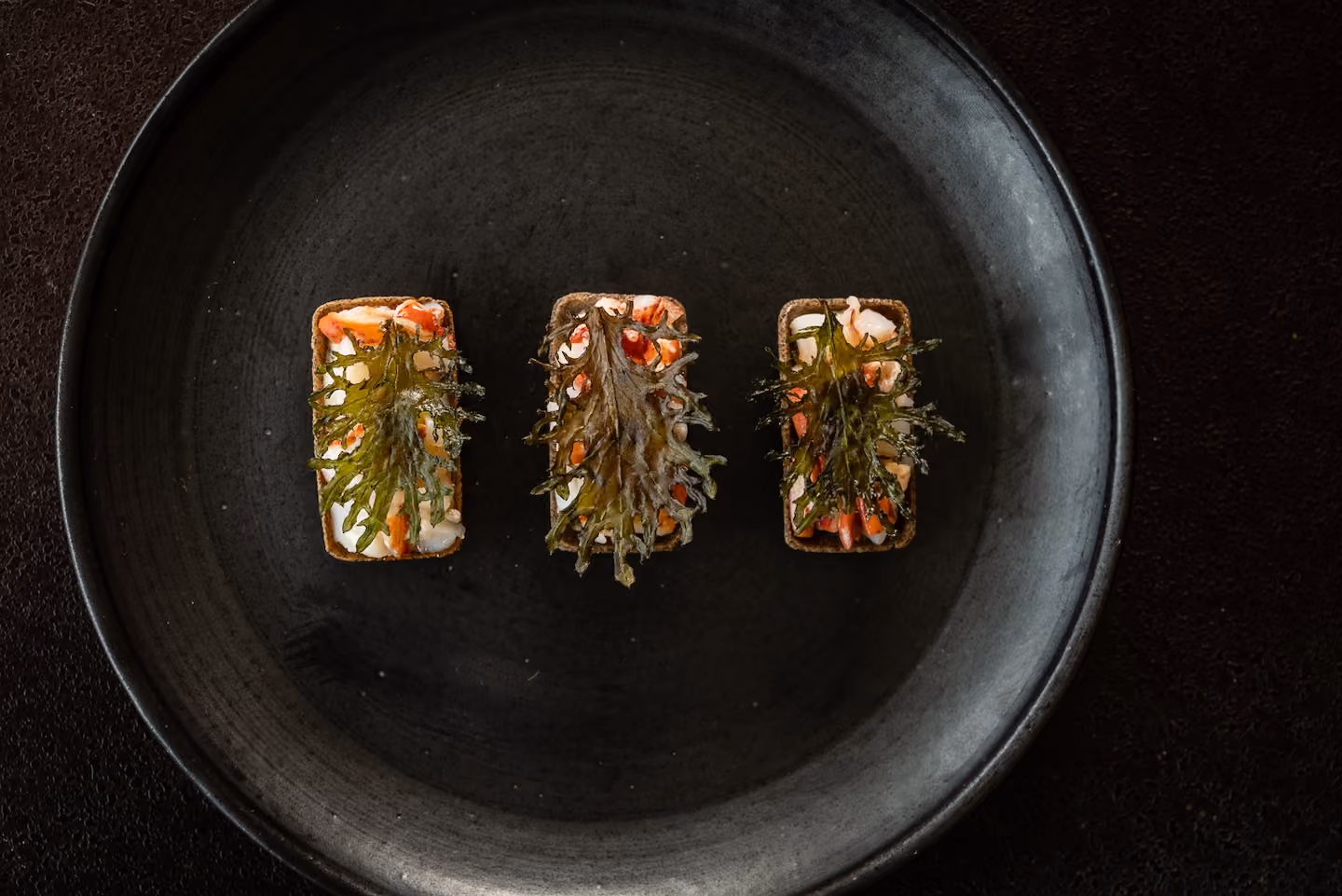Row of tartlets topped with crab and crispy seaweed on a dark plate