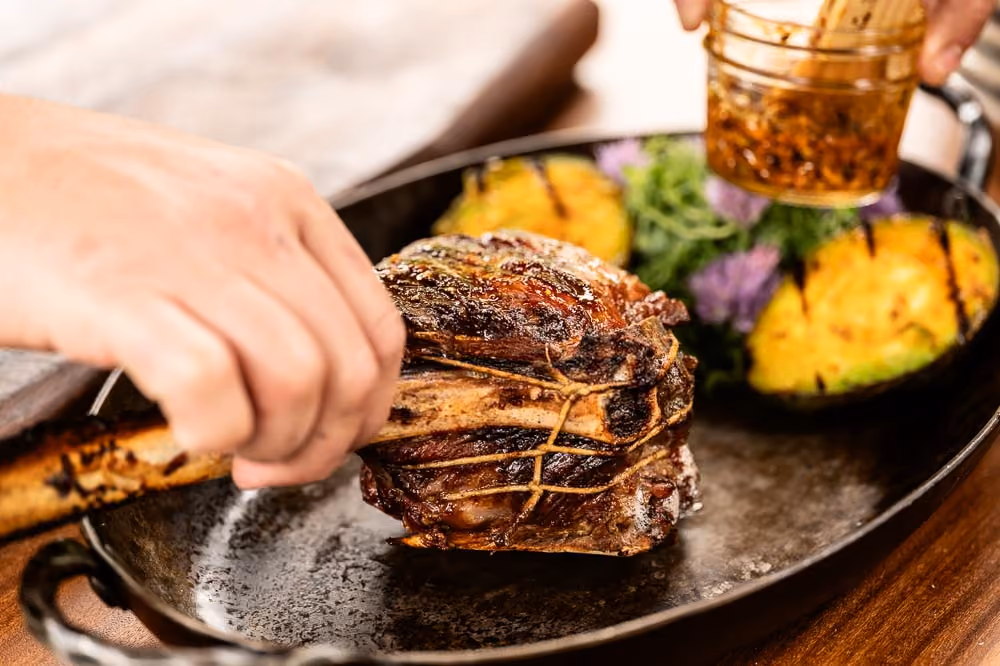 Chef basting a tied roast beside grilled avocado halves in a cast iron pan