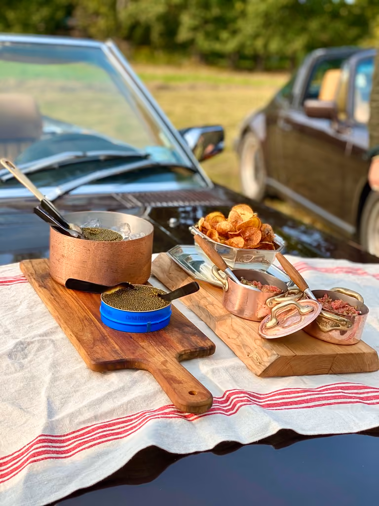 Caviar tins and fried potato chips served on wooden boards beside a vintage car