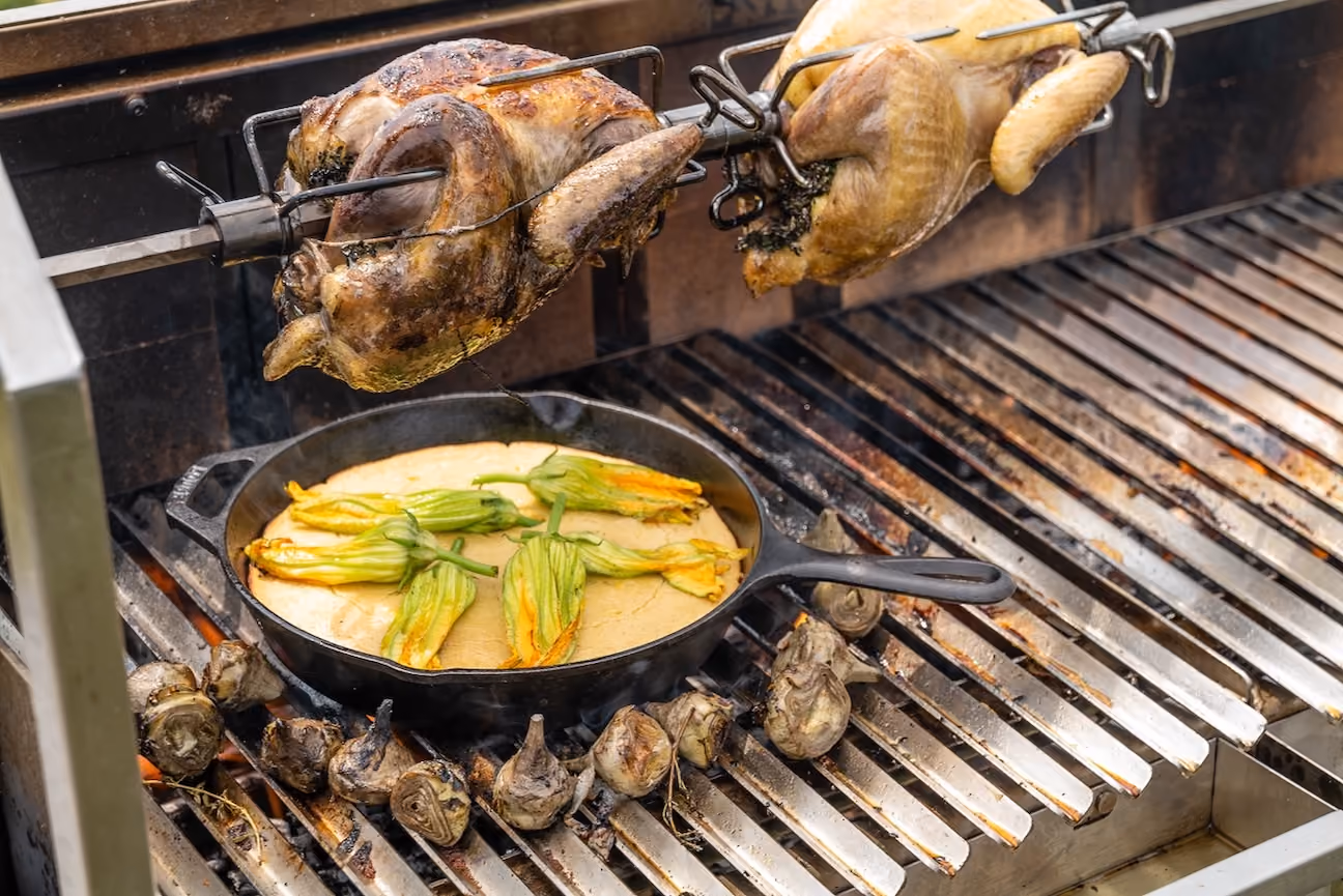 Two whole chickens on a rotisserie grilling above a skillet of squash blossoms