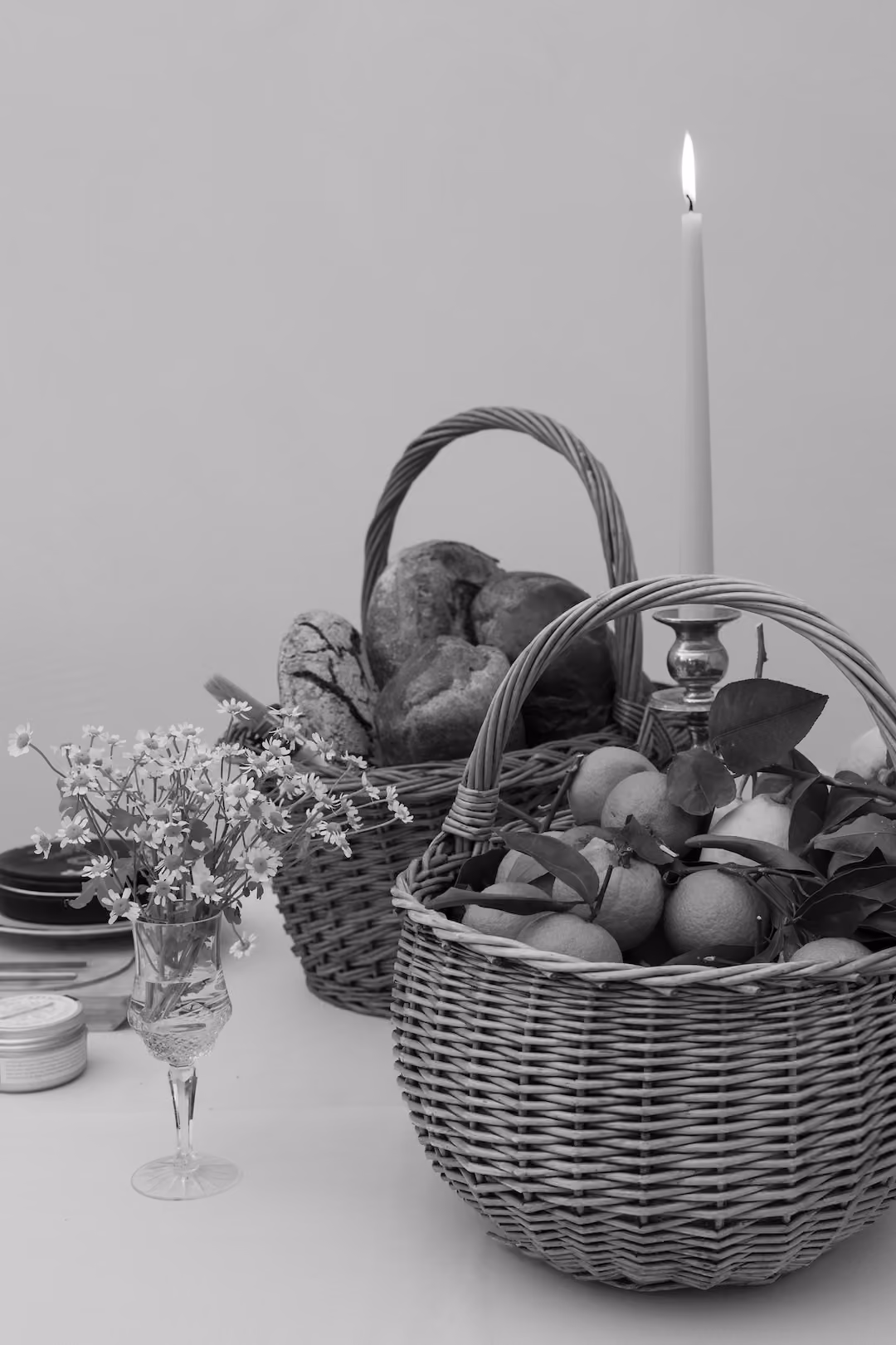 Black-and-white still life of baskets with bread and citrus, a lit candle, and a wine glass