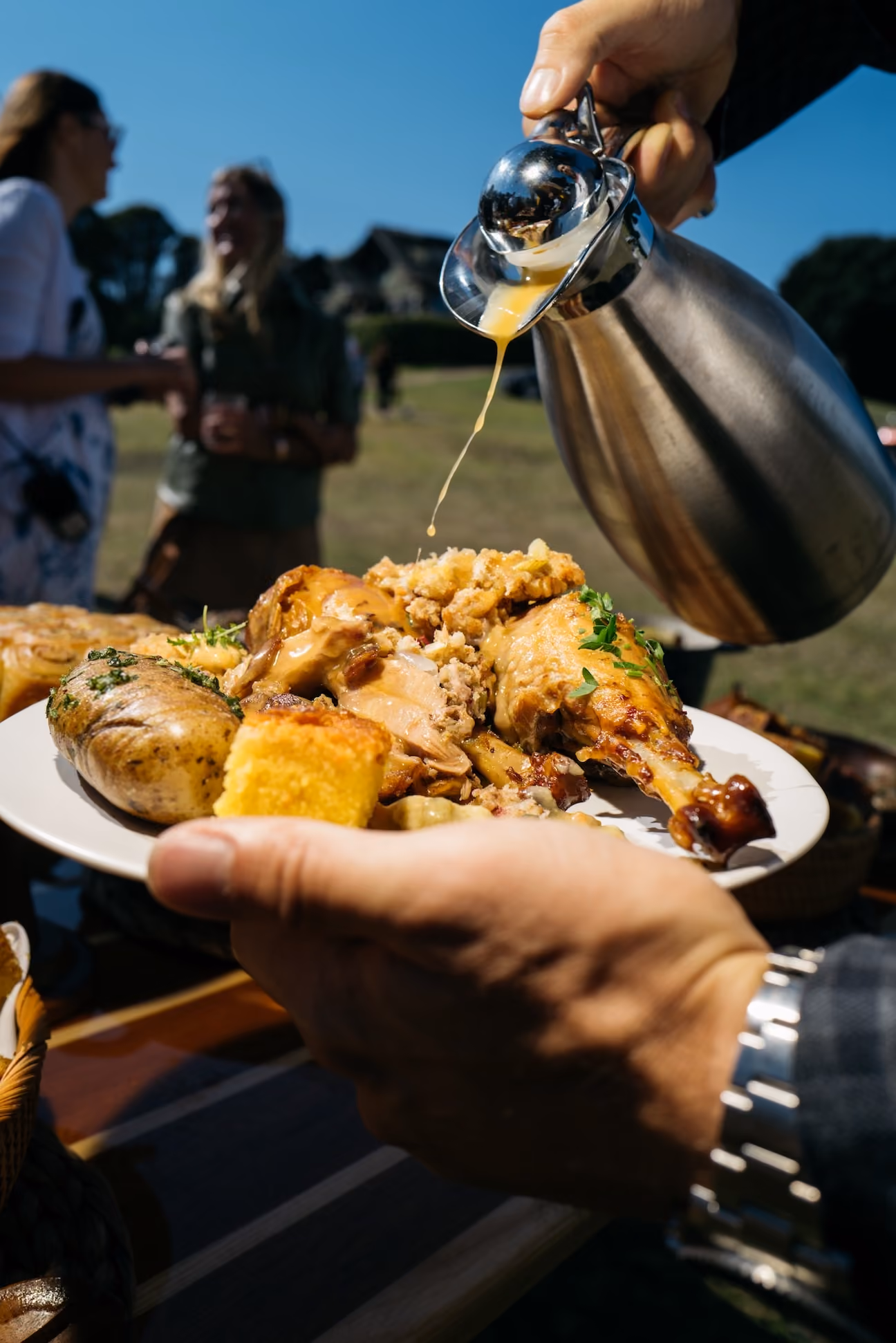 Person pouring gravy over a plate of roasted chicken and sides outdoors