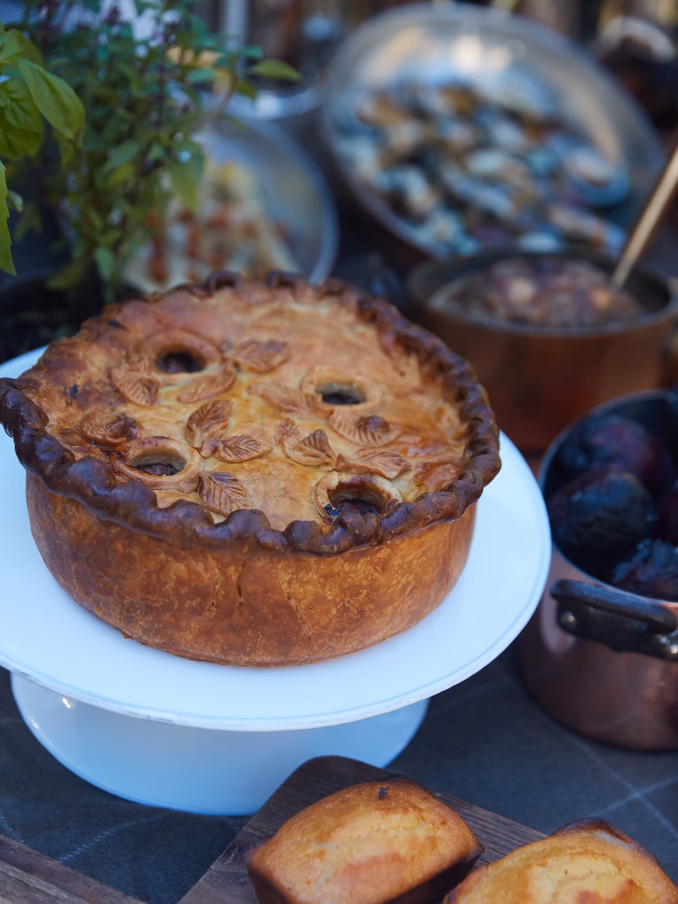 Golden baked meat pie on a cake stand beside cornbread squares at an outdoor table setting