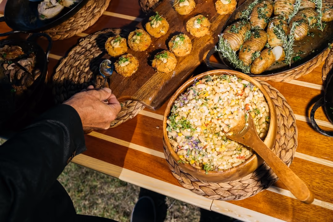 Overhead outdoor buffet spread with croquettes, bean salad, and roasted potatoes as a hand serves food