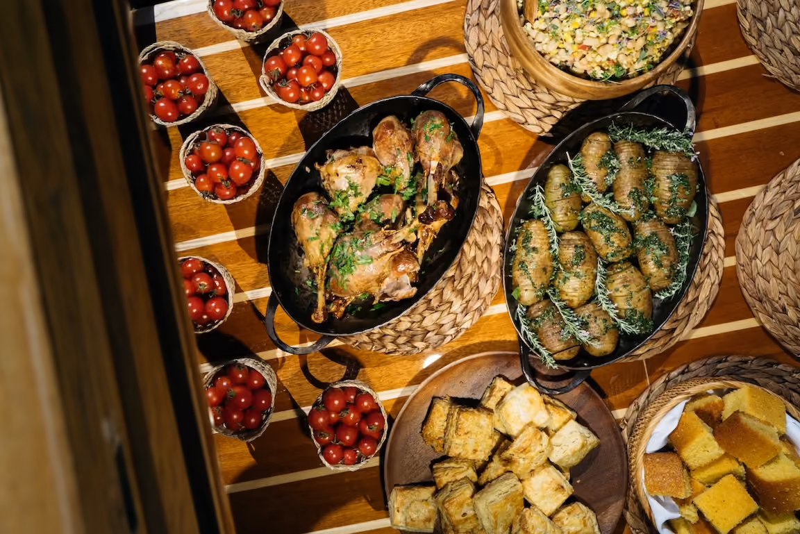 Overhead outdoor spread with roasted duck, hasselback potatoes, cherry tomatoes, biscuits, and cornbread