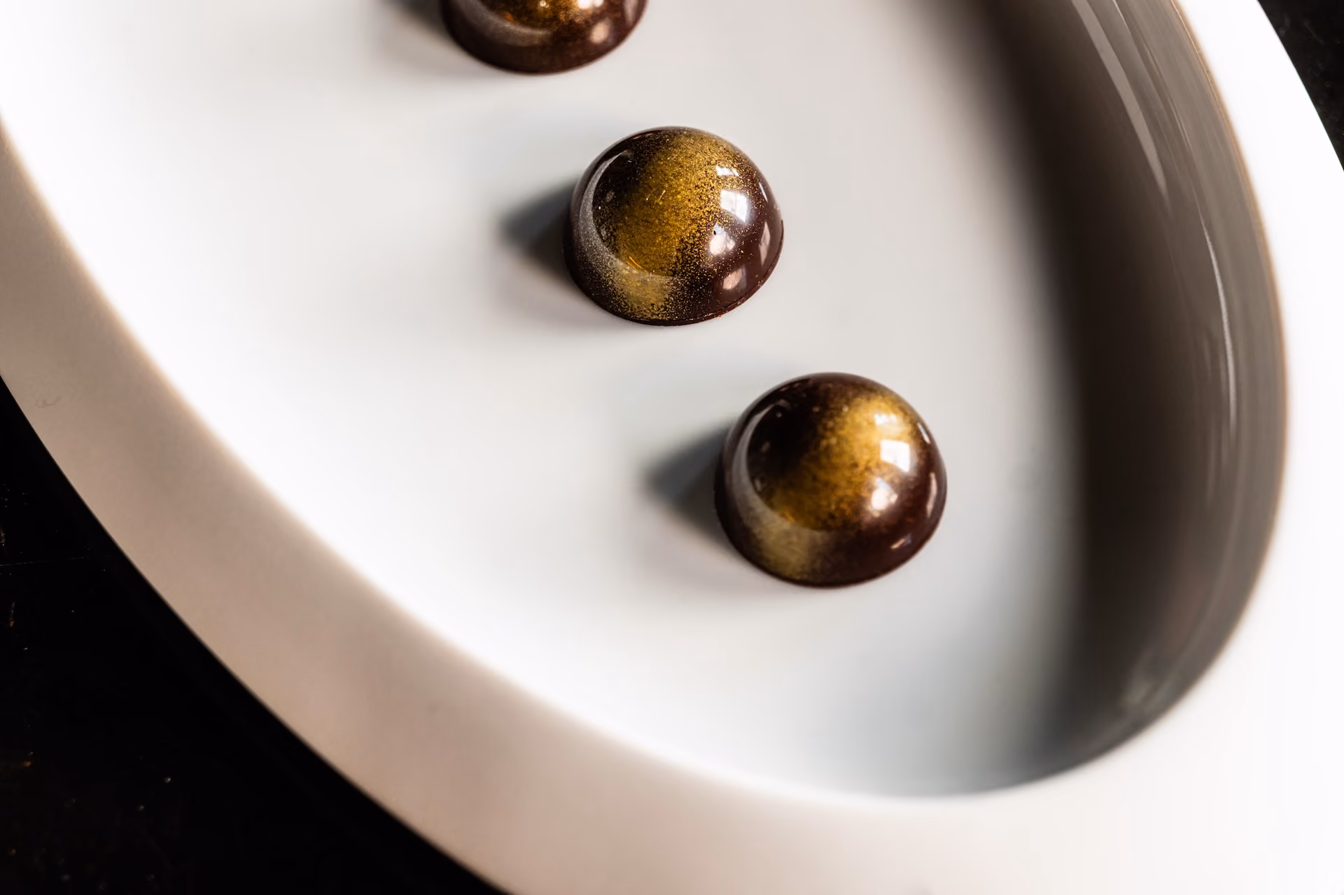 Dark chocolate dome bonbons with gold dust arranged in a white oval dish