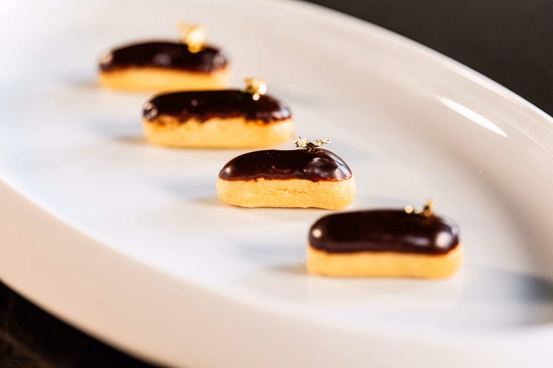 Chocolate-glazed eclairs finished with gold leaf, lined up on a white plate