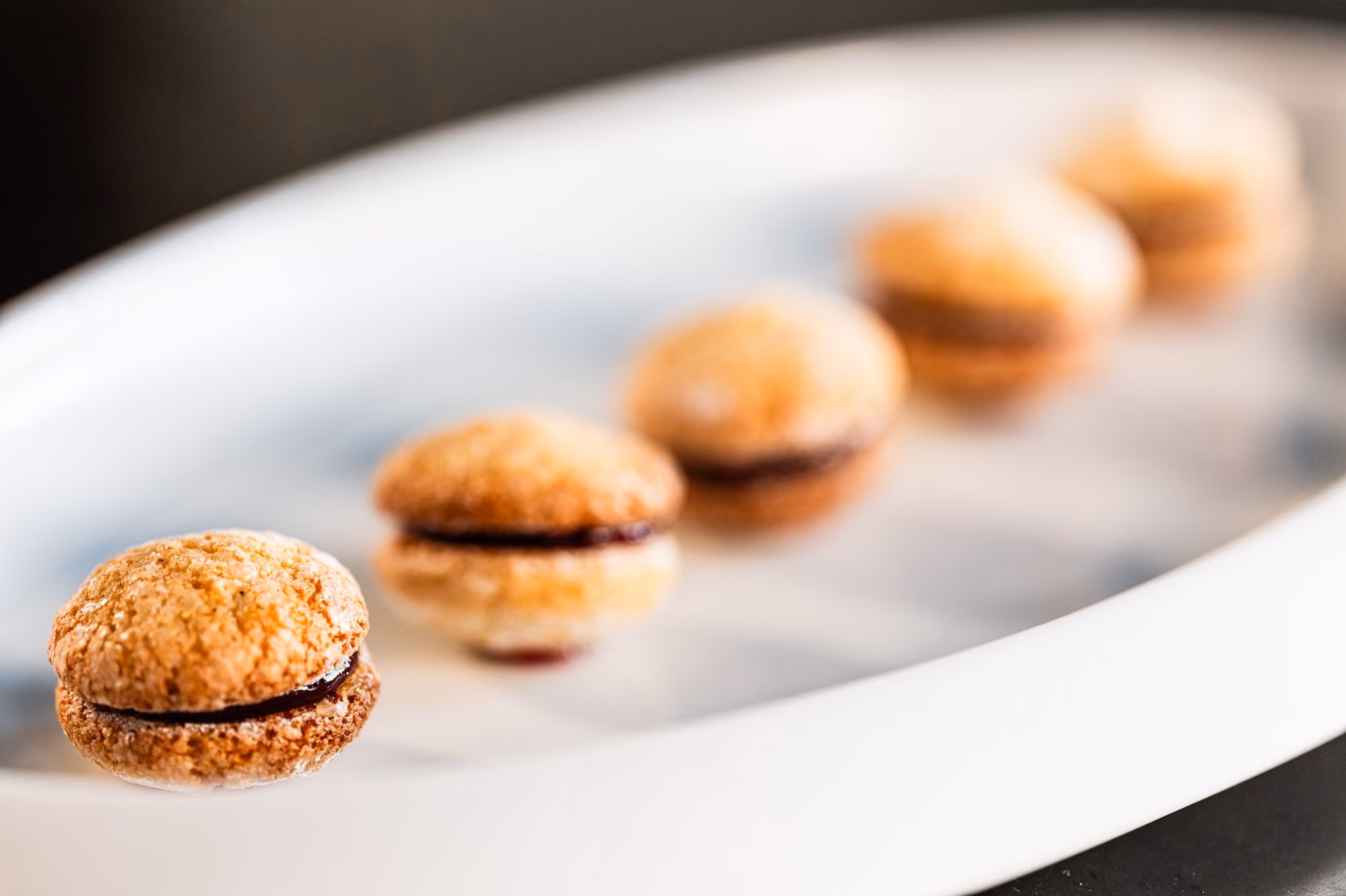 Five sugar-dusted sandwich cookies filled with dark jam, lined up on a white plate