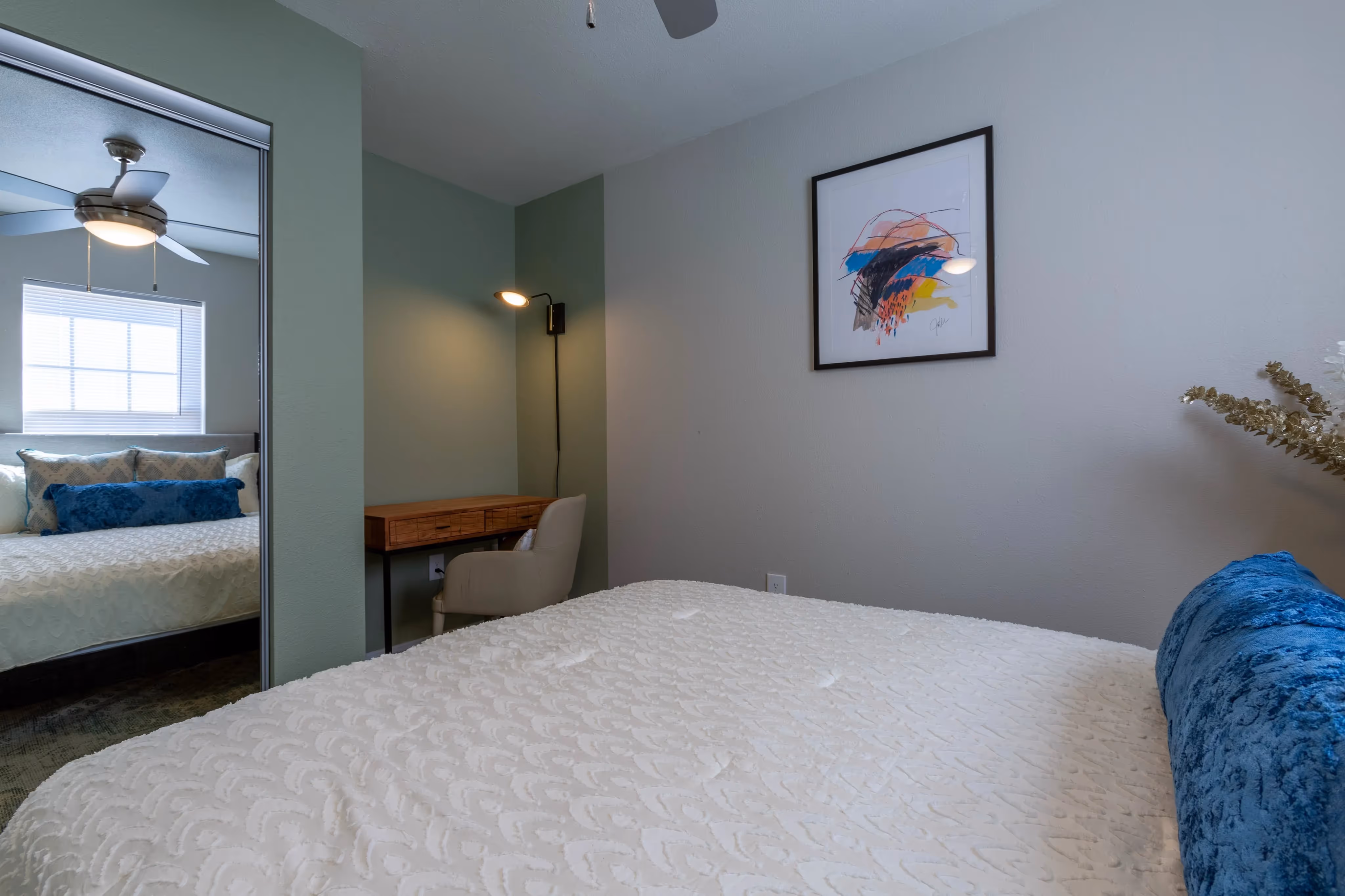 Bedroom with a mirrored closet door, a bed with blue accent pillows, and a desk, in an apartment at Ascension at Ironwood in Tucson, AZ.