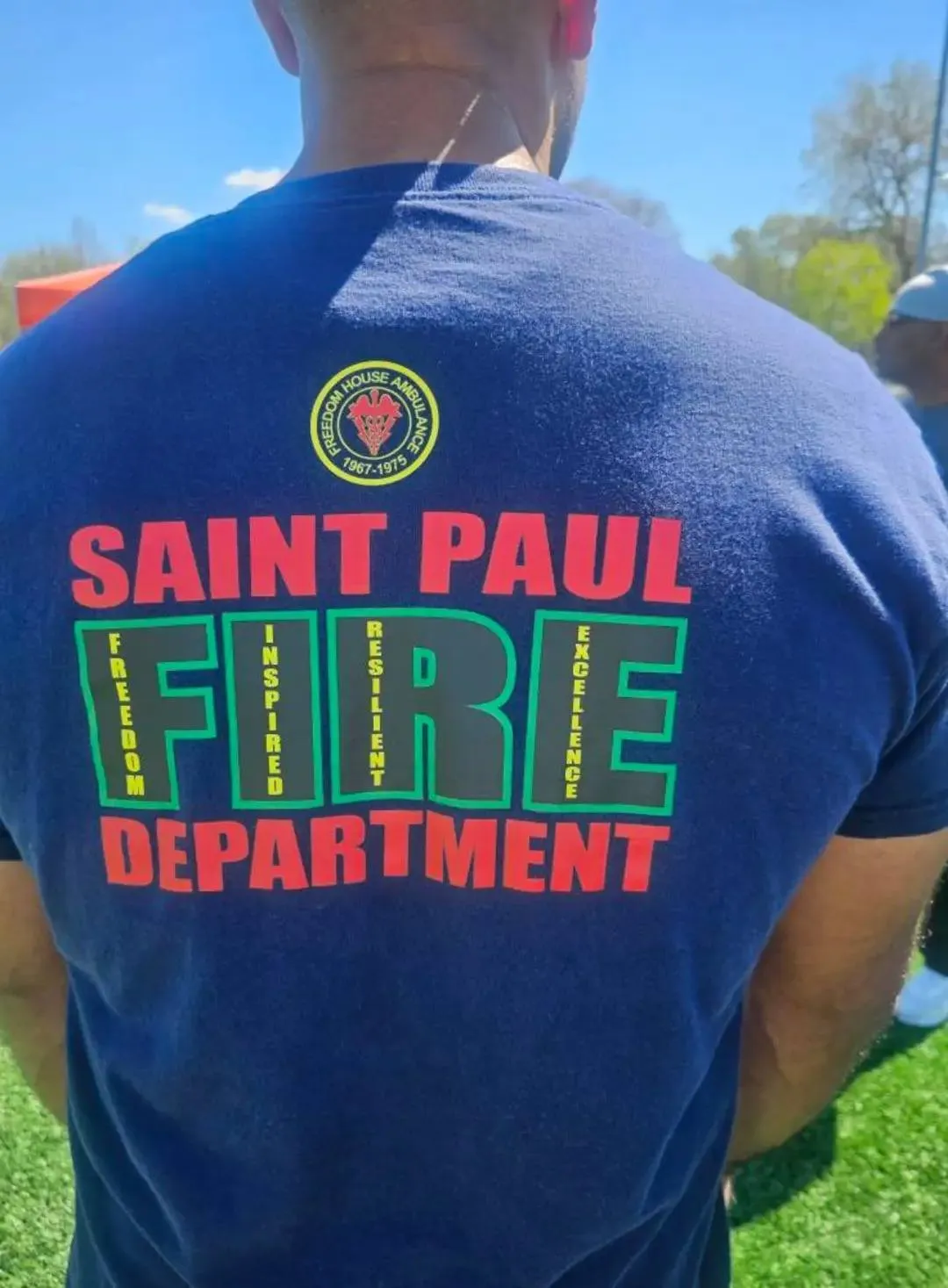 Back view of person wearing a navy blue T-shirt with Saint Paul Fire Department logo and words Freedom, Inspired, Resilient, Excellence on the back.
