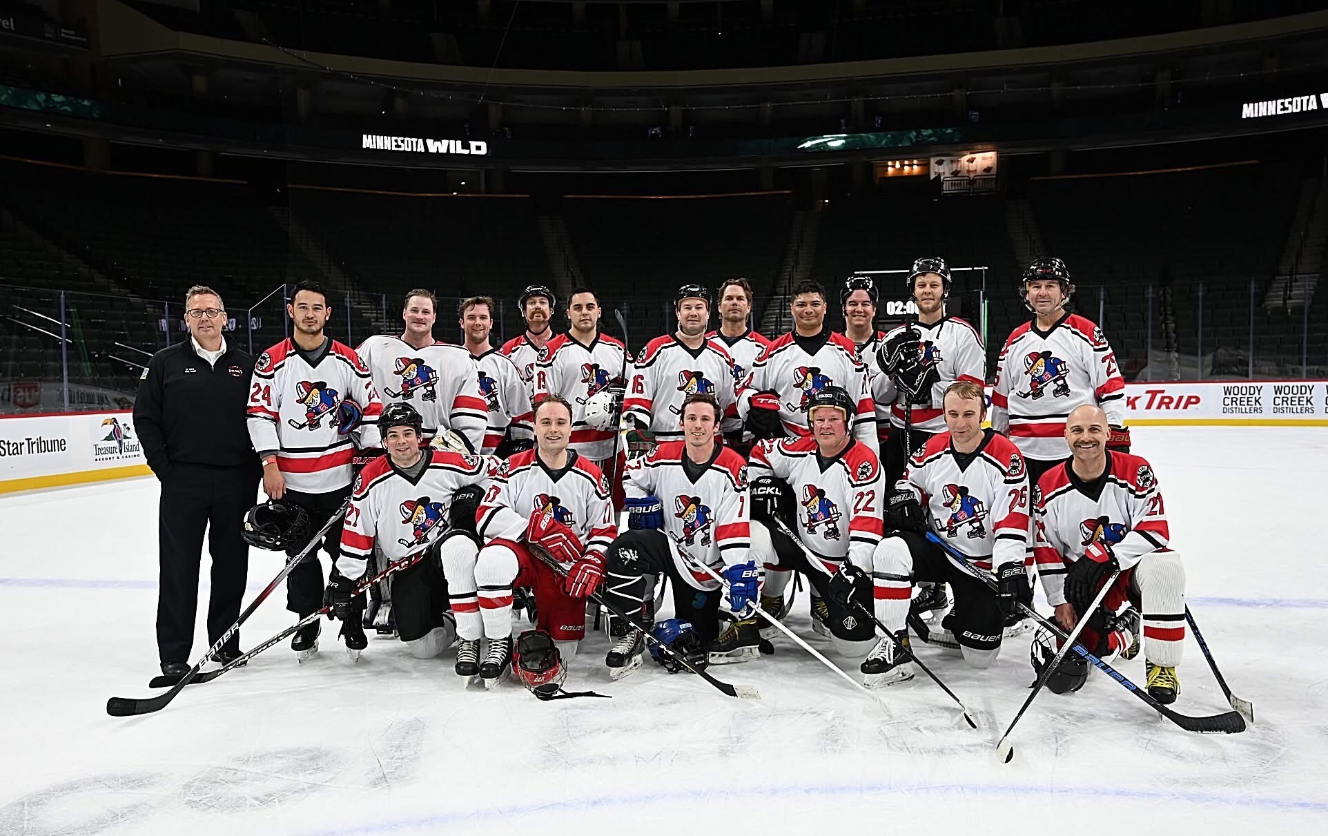 Group photo of an adult ice hockey team wearing white, red, and black jerseys on an ice rink with Minnesota Wild signage in the background.
