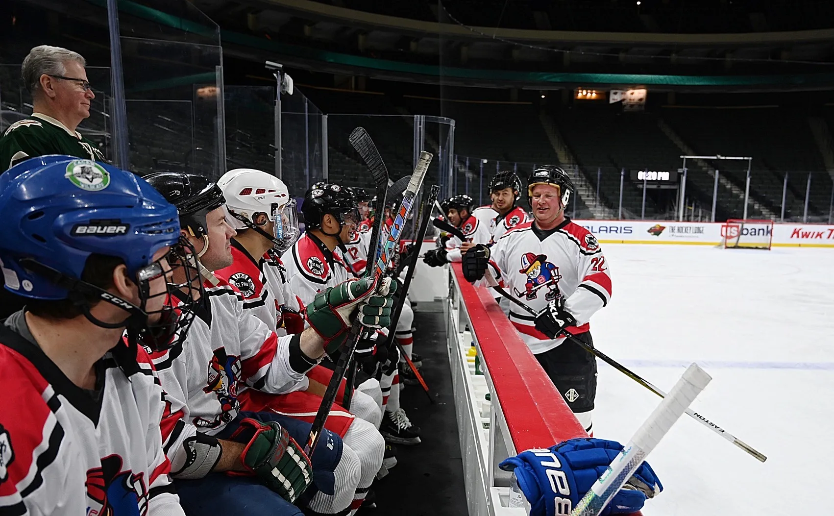 Ice hockey players in white and red jerseys sitting on the bench, with one player standing on the ice holding a hockey stick.