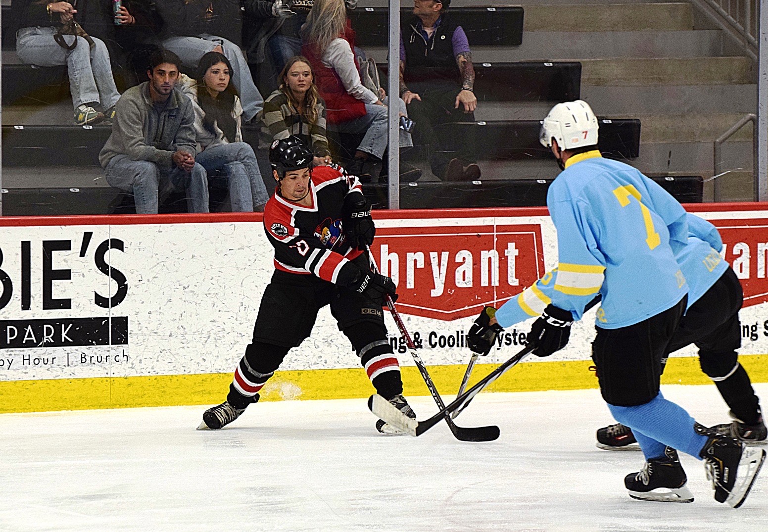 Hockey player in black and red jersey shooting the puck while being challenged by two players in light blue jerseys on an ice rink.