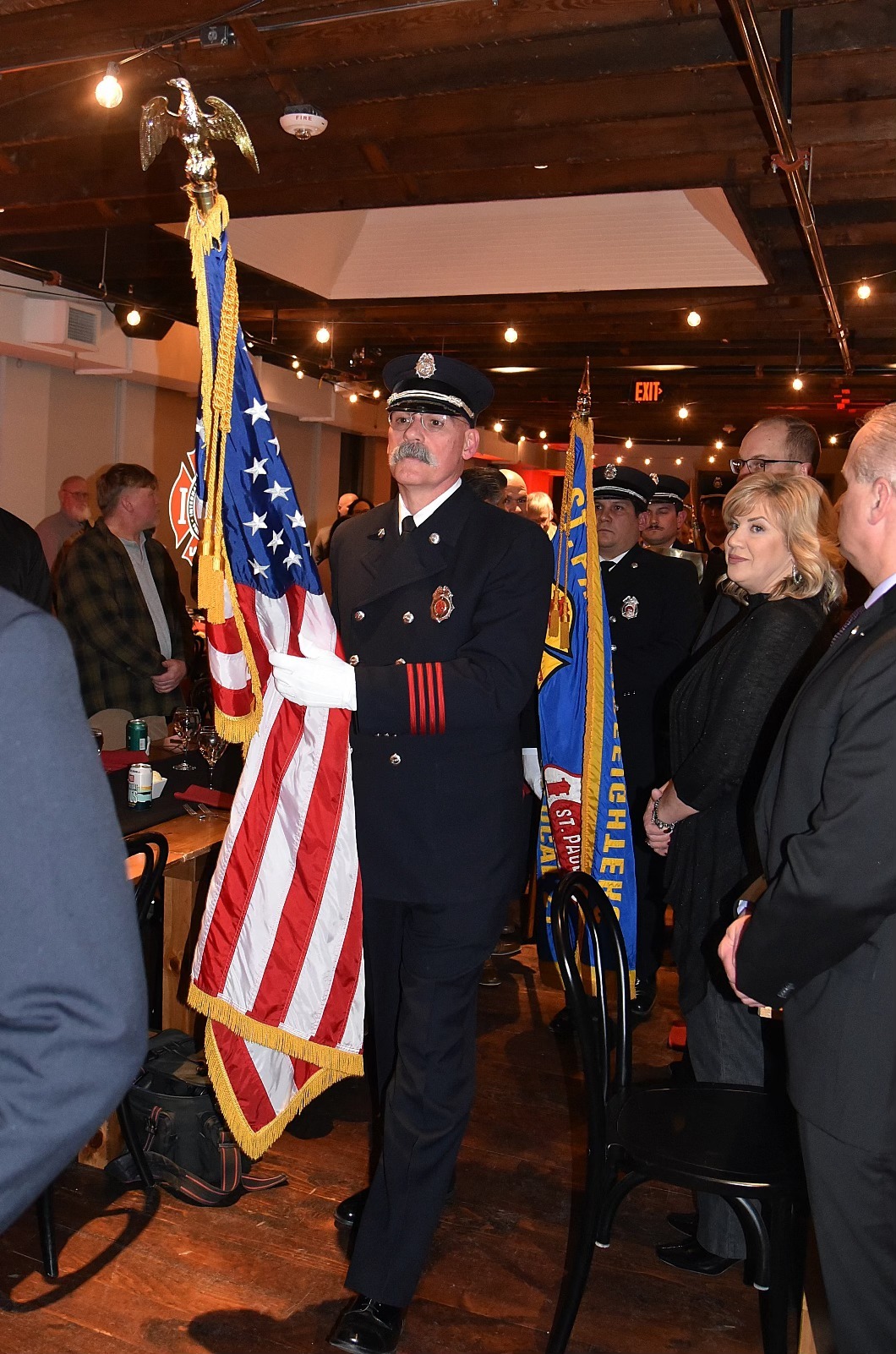 Uniformed firefighter carrying the American flag during a formal indoor ceremony with attendees standing respectfully.