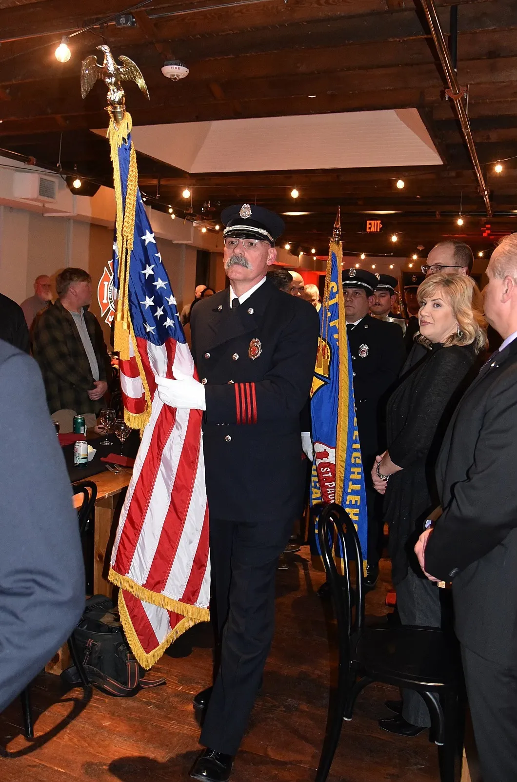 Uniformed firefighter carrying the American flag during a formal indoor ceremony with attendees standing respectfully.