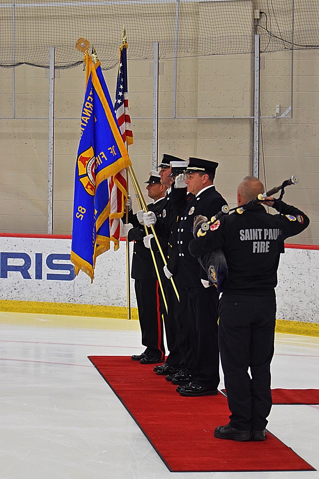 Four firefighters in uniform standing on a red carpet on an ice rink, three holding flags and one playing bagpipes, during a ceremony.