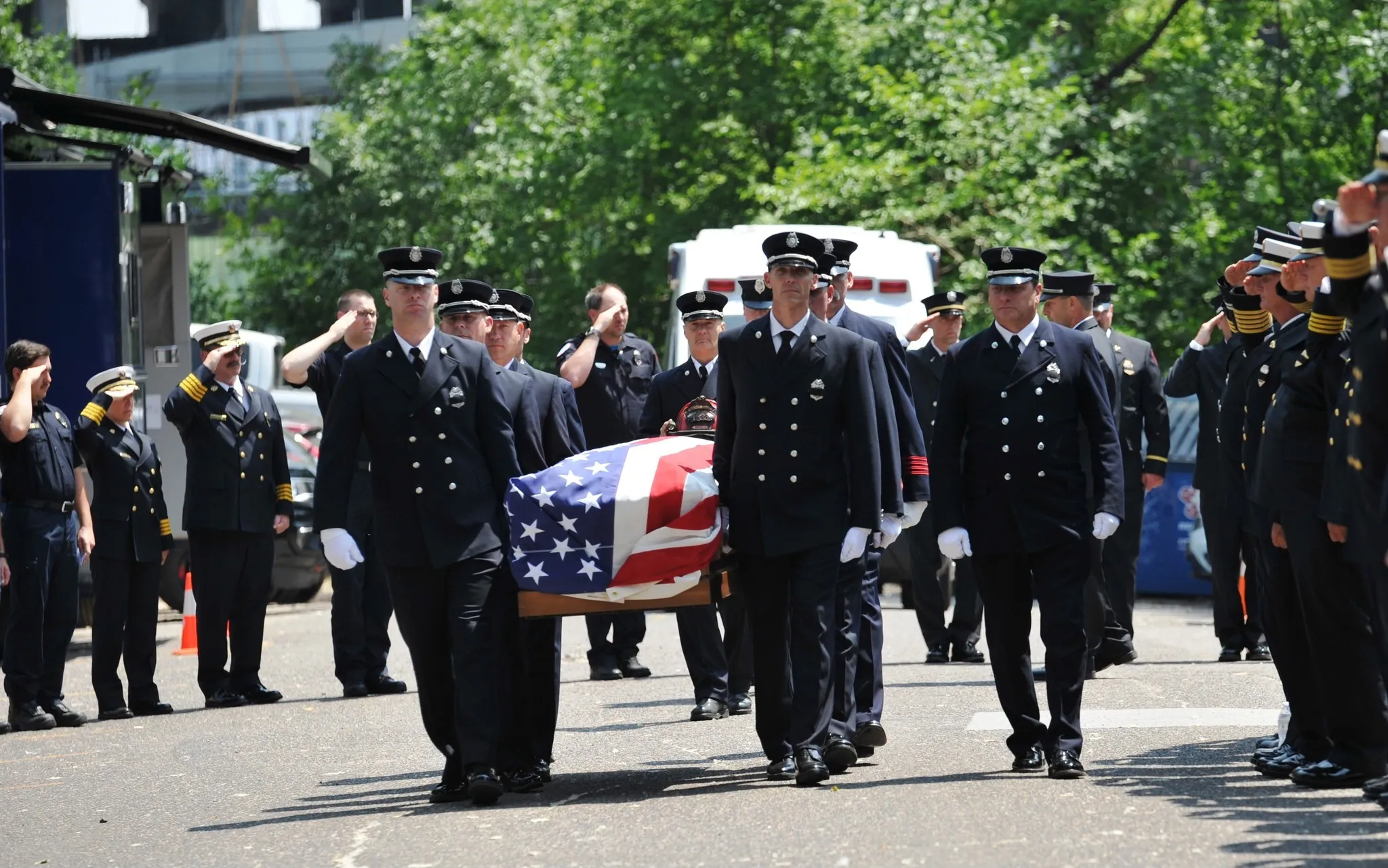 Uniformed firefighters solemnly carry a flag-draped casket during a funeral procession while others salute on both sides of the street.