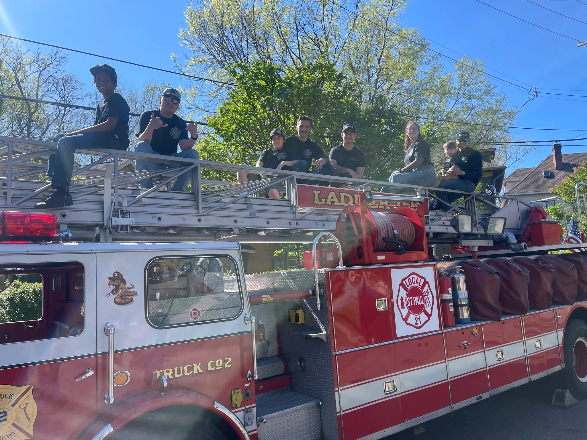 Seven people, including children and adults, sitting on an extended ladder of a red fire truck labeled Local St. Paul, Truck Co 2.