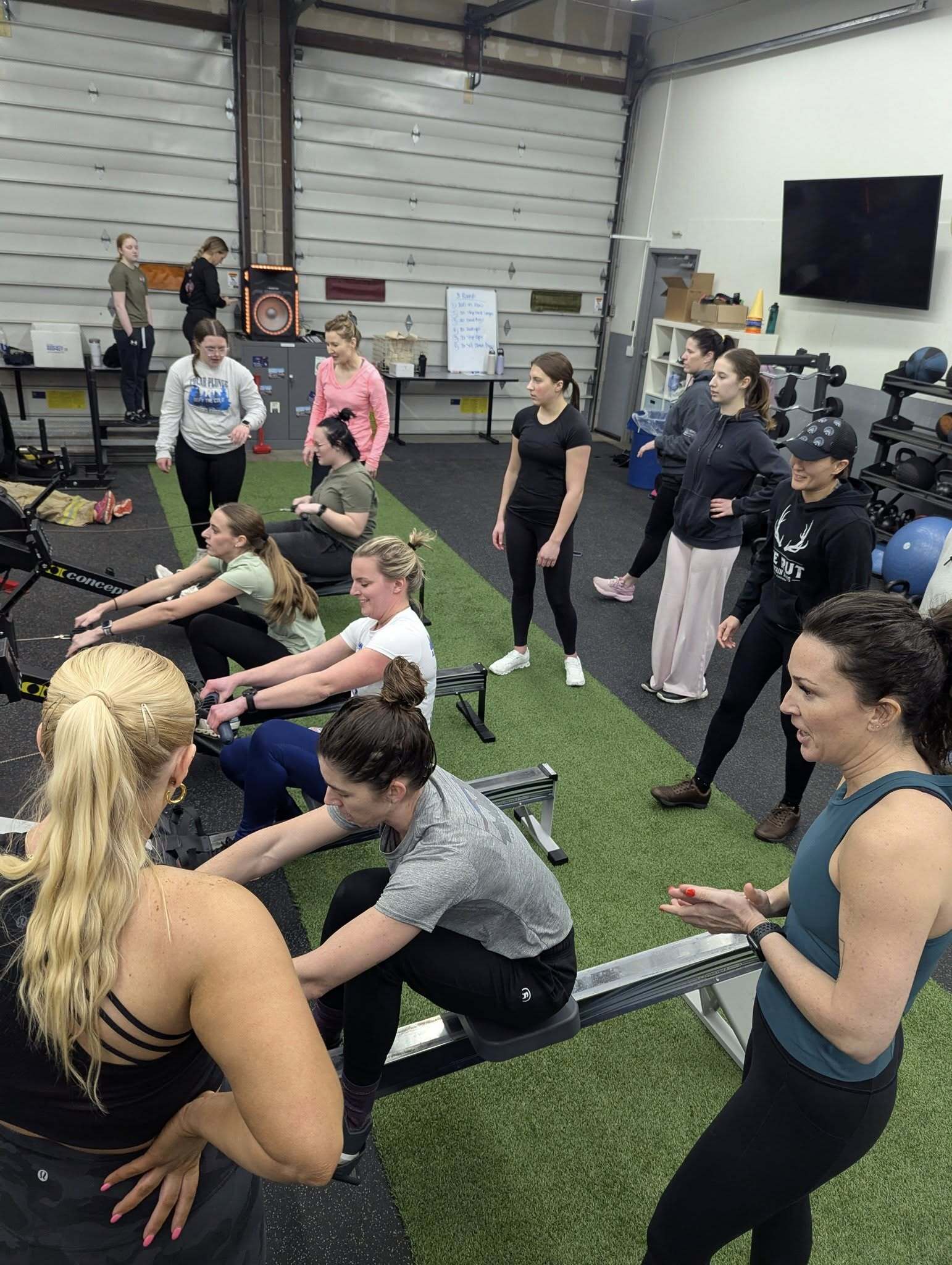 Group of women working out on rowing machines indoors while others stand nearby in a gym with green turf flooring.