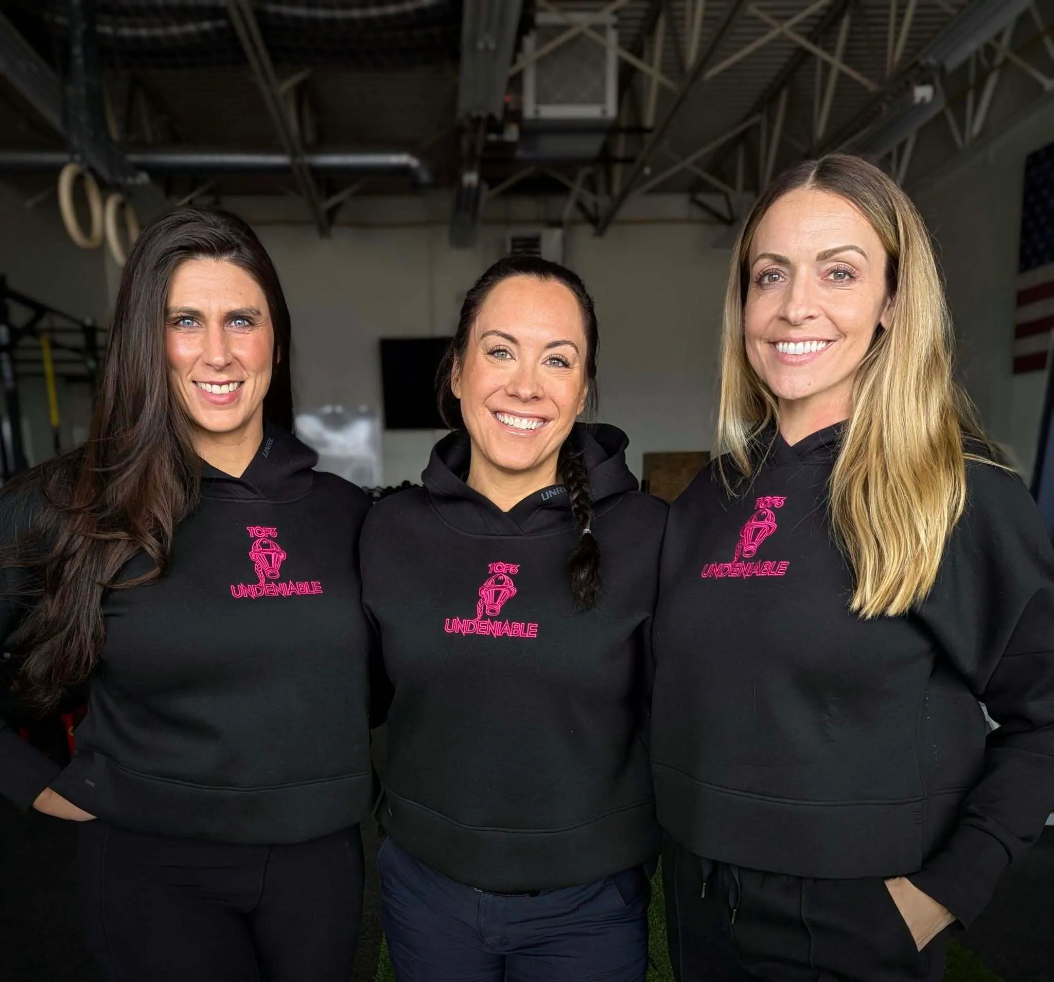 Three smiling women standing side by side wearing black hoodies with pink 'TCF3 Undeniable' logos in a gym setting.
