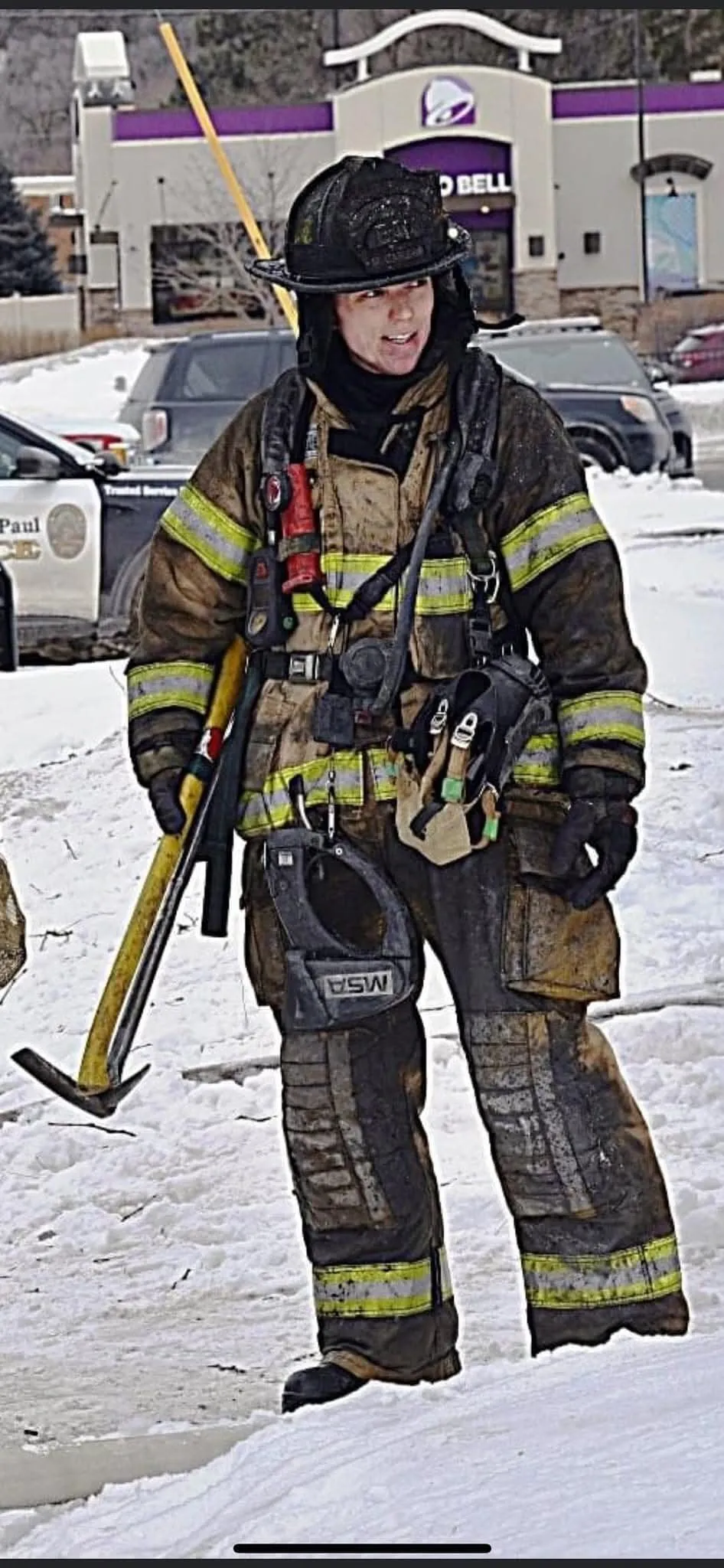 Firefighter in full gear holding a tool, standing on snow with police cars and a Taco Bell restaurant in the background.