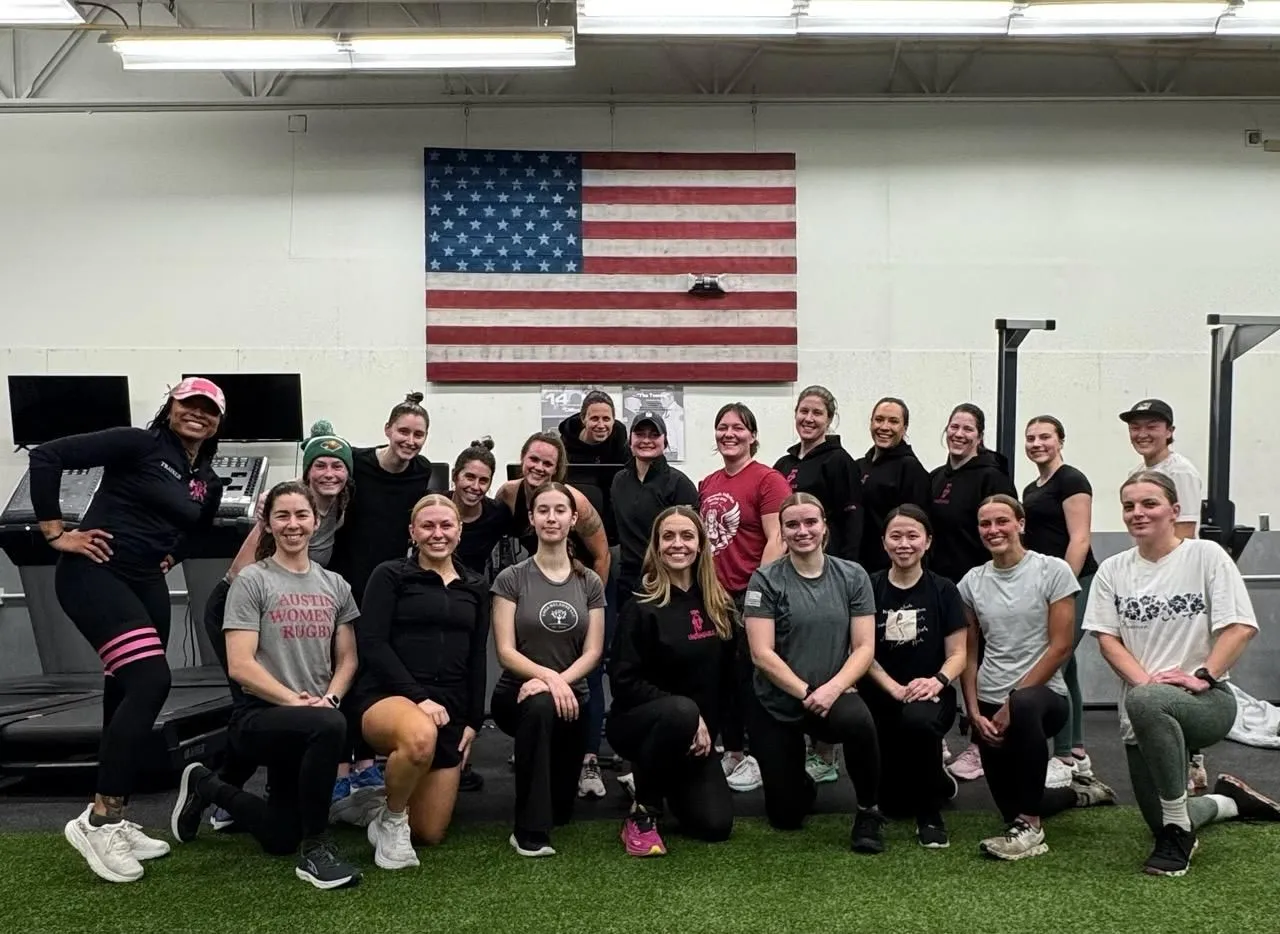Group of 20 women posing together inside a gym with an American flag on the wall behind them.