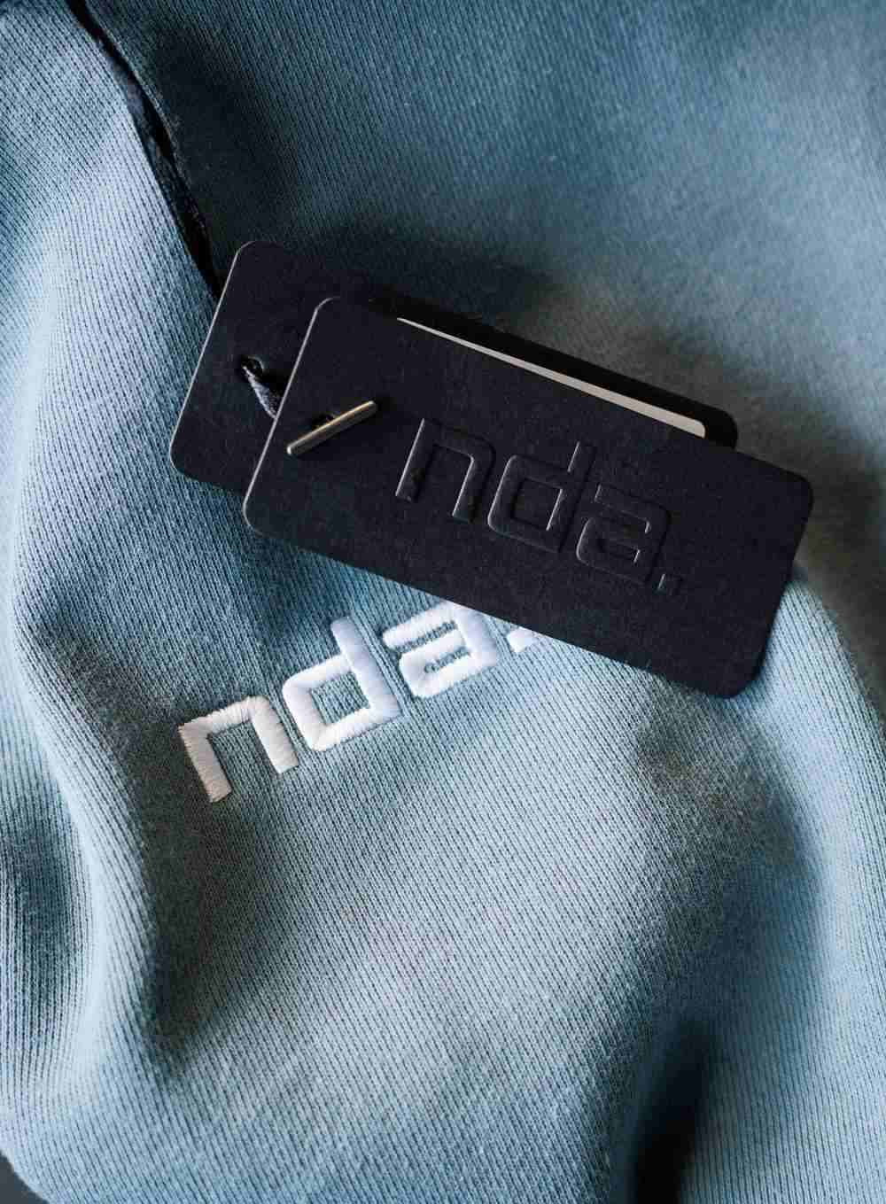 A blue NDA hoodie with the clothing tag laying next to the logo.