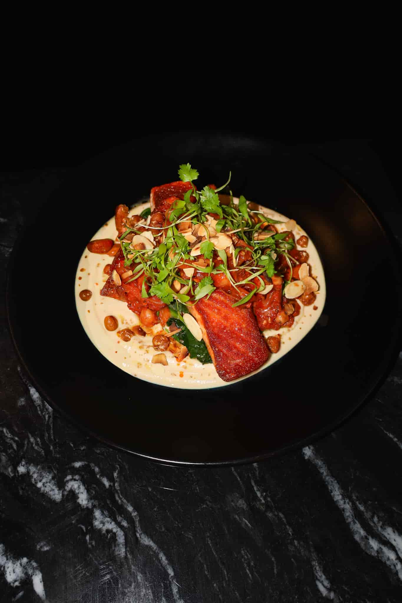 Plated dish with seared salmon fillets on creamy sauce, topped with sliced almonds and fresh herbs, served on a black plate.