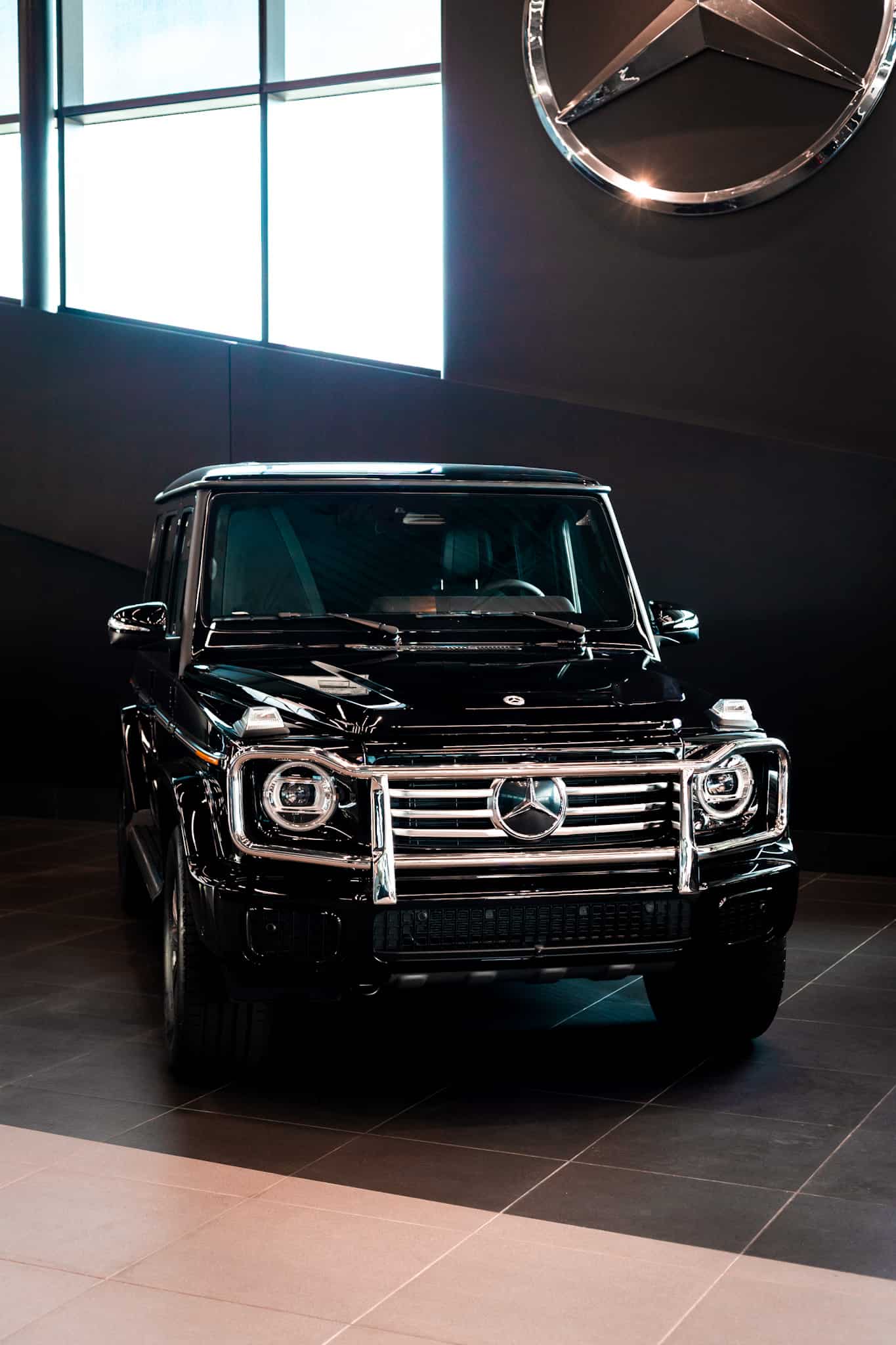 Front view of a black Mercedes-Benz G-Class SUV in a showroom with large windows and the Mercedes emblem on the wall.
