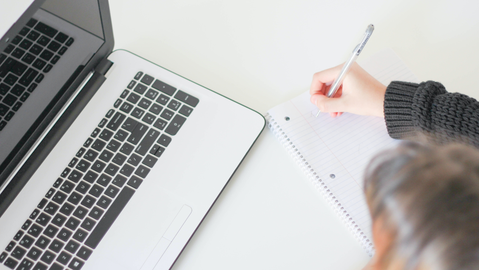 Person writing in notebook next to laptop with knitted sleeve