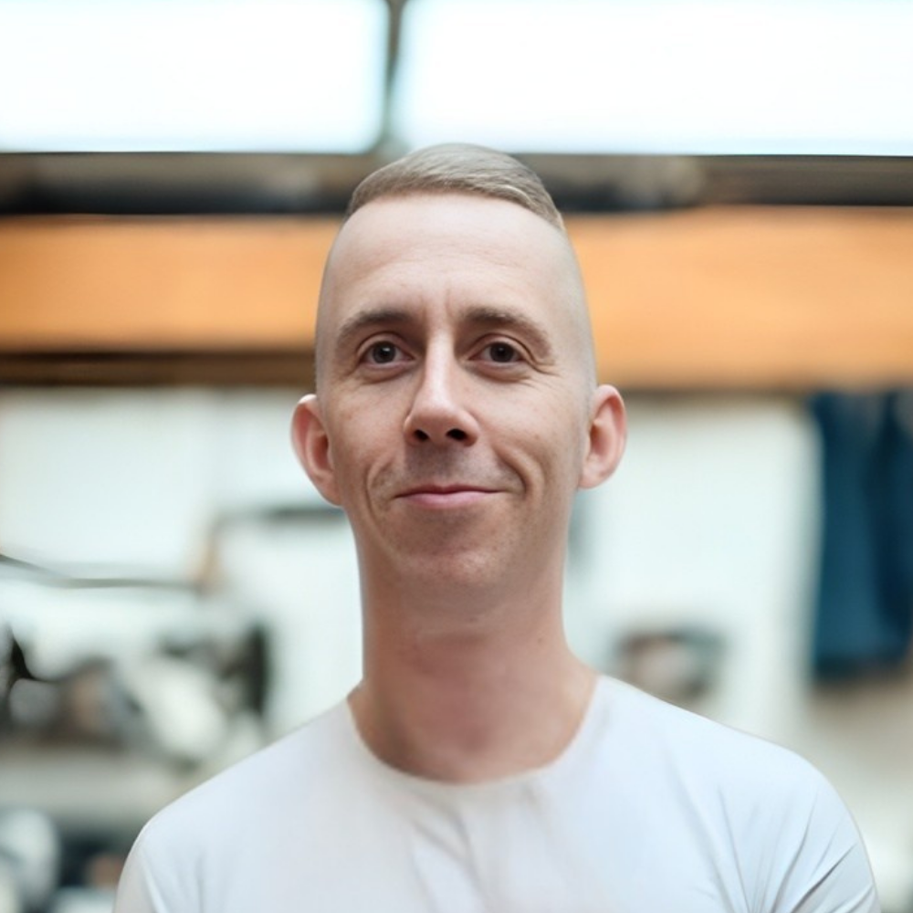 Mężczyzna z krótkimi włosami i białą koszulką, uśmiechający się na rozmytym tle wnętrza.