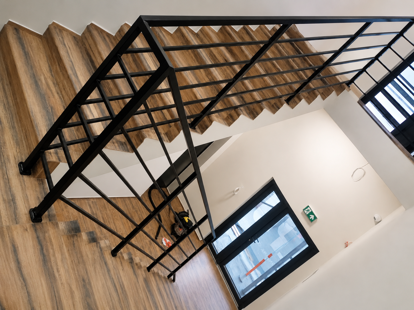 Nowoczesne schody z drewnianymi stopniami i czarną metalową balustradą w minimalistycznym wnętrzu z betonową ścianą i dużymi oknami.