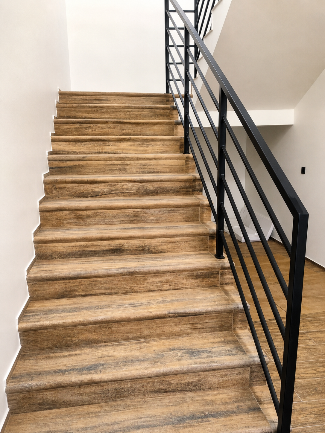 Nowoczesne schody z drewnianymi stopniami i czarną metalową balustradą w minimalistycznym wnętrzu z betonową ścianą i dużymi oknami.